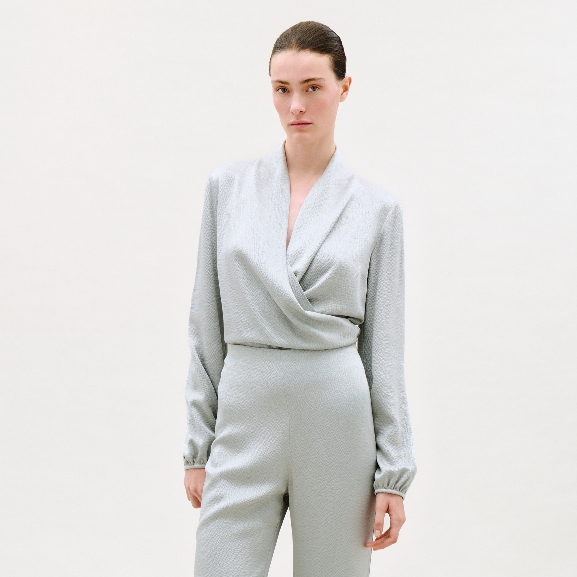 A woman wearing the Tea Blouse in light gray with long sleeves and wrap style, paired with matching high-waisted pants, stands against a plain light background, gazing confidently at the camera.