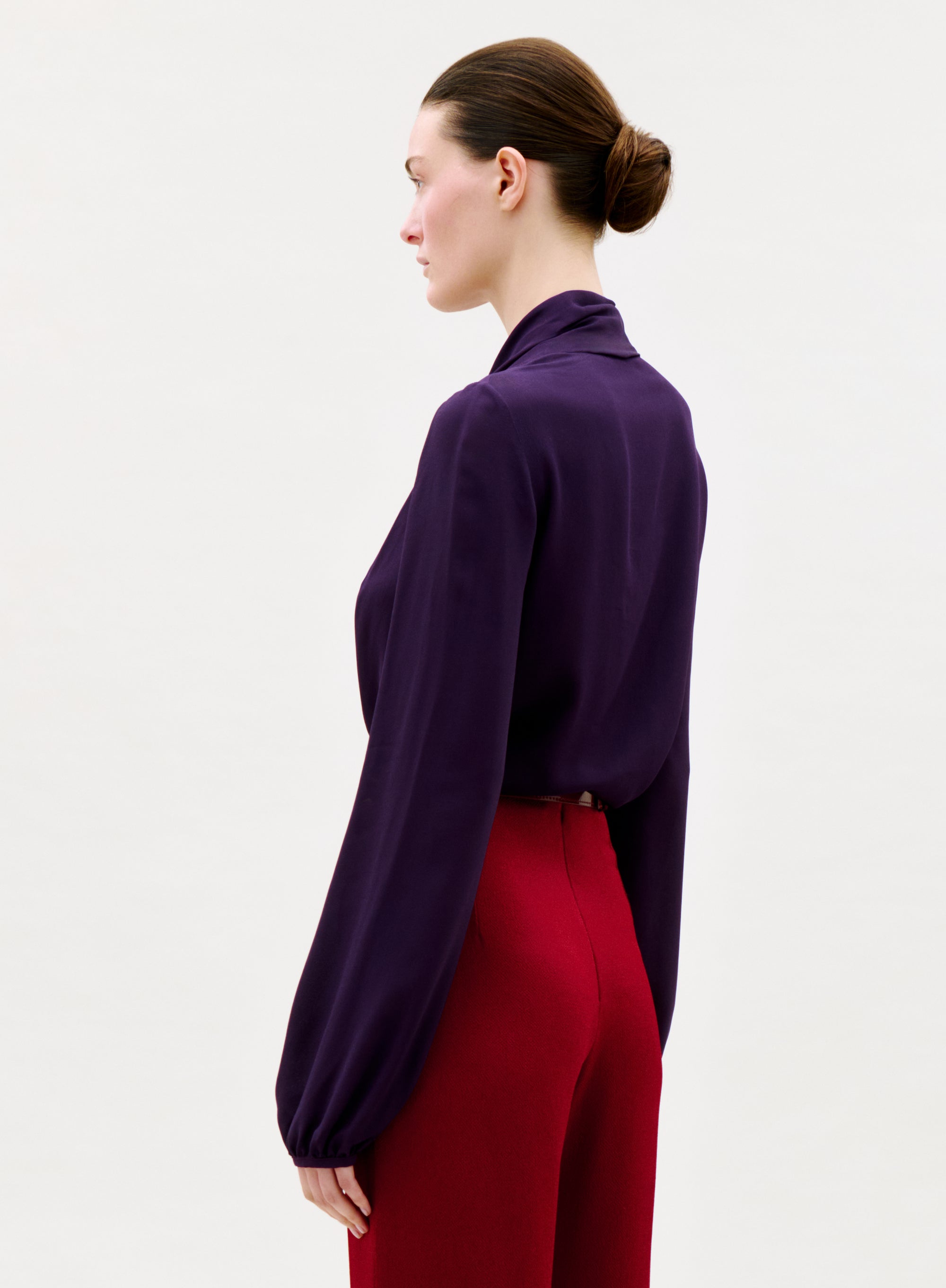 A woman stands in profile against a plain light background, wearing the Tea Blouse—long-sleeved and dark purple—with high-waisted red pants. Her brown hair is neatly tied back in a low bun.