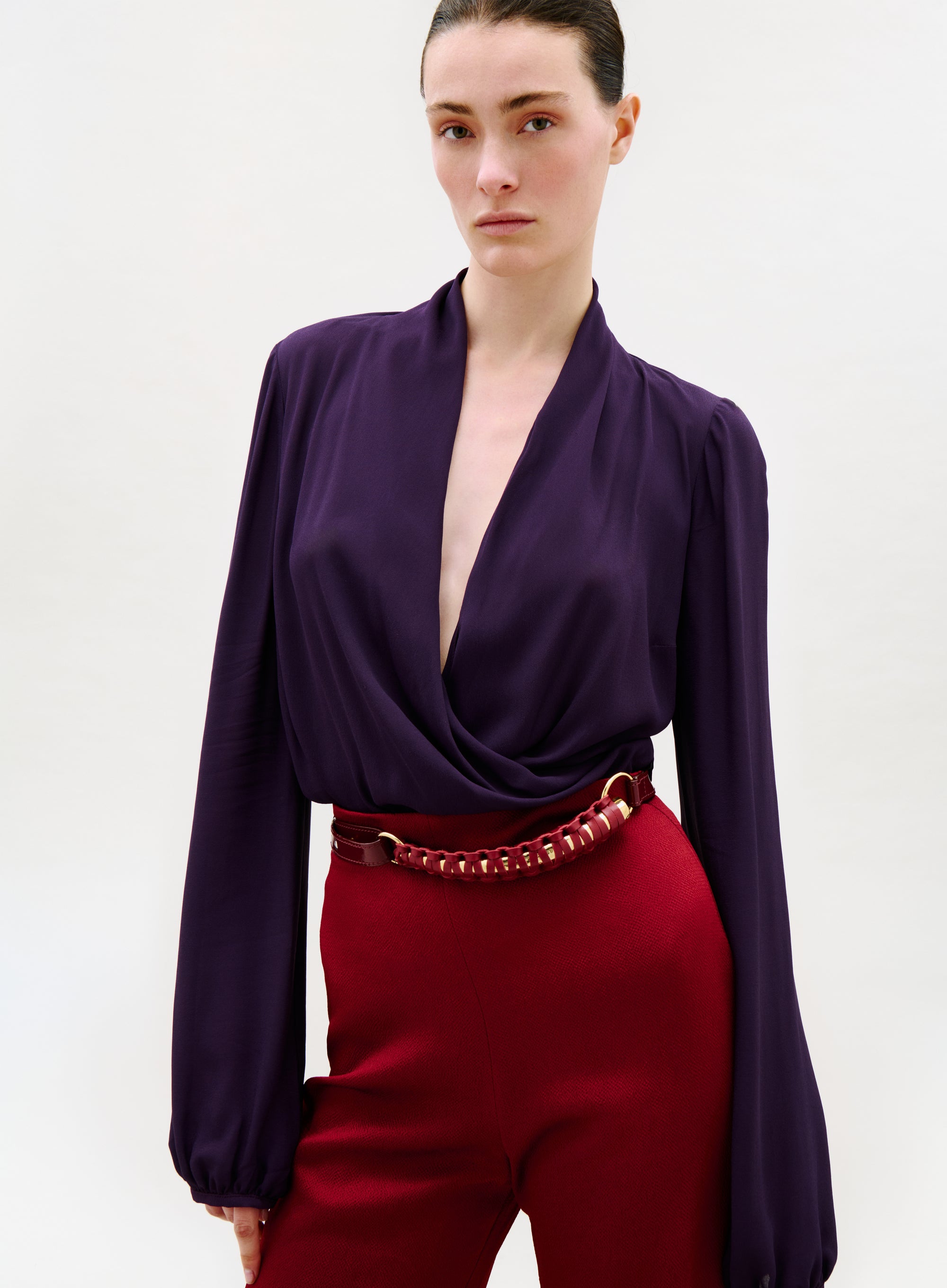 A person with fair skin and dark hair pulled back wears the deep purple, long-sleeved Tea Blouse with high-waisted red pants and a wide belt, standing against a plain light background.