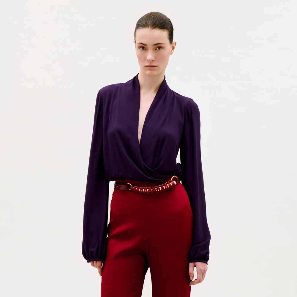 A person with brown hair pulled back stands against a plain light background, wearing the deep purple Tea Blouse with long sleeves and draped details, paired with high-waisted red trousers with a decorative belt.