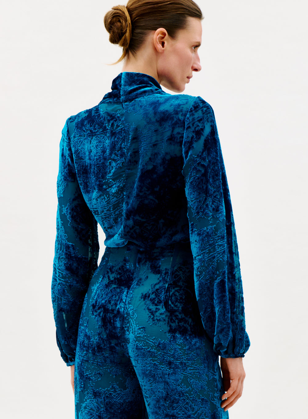 A woman with light skin and brown hair in a bun is seen from the back, wearing the Tea Blouse in deep blue Italian velvet with long sleeves, standing against a plain white background.