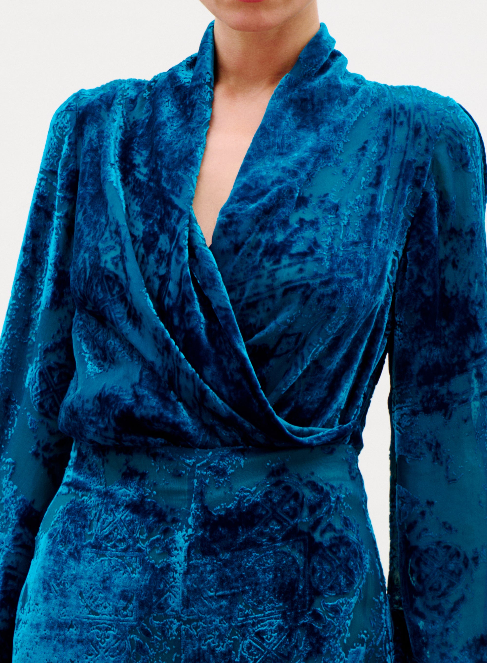A person wearing the Tea Blouse, made from luxurious Italian blue crushed velvet with long sleeves and a deep V-neckline. The blouse has a soft, textured look with intricate patterns, set against a plain, light-colored background.