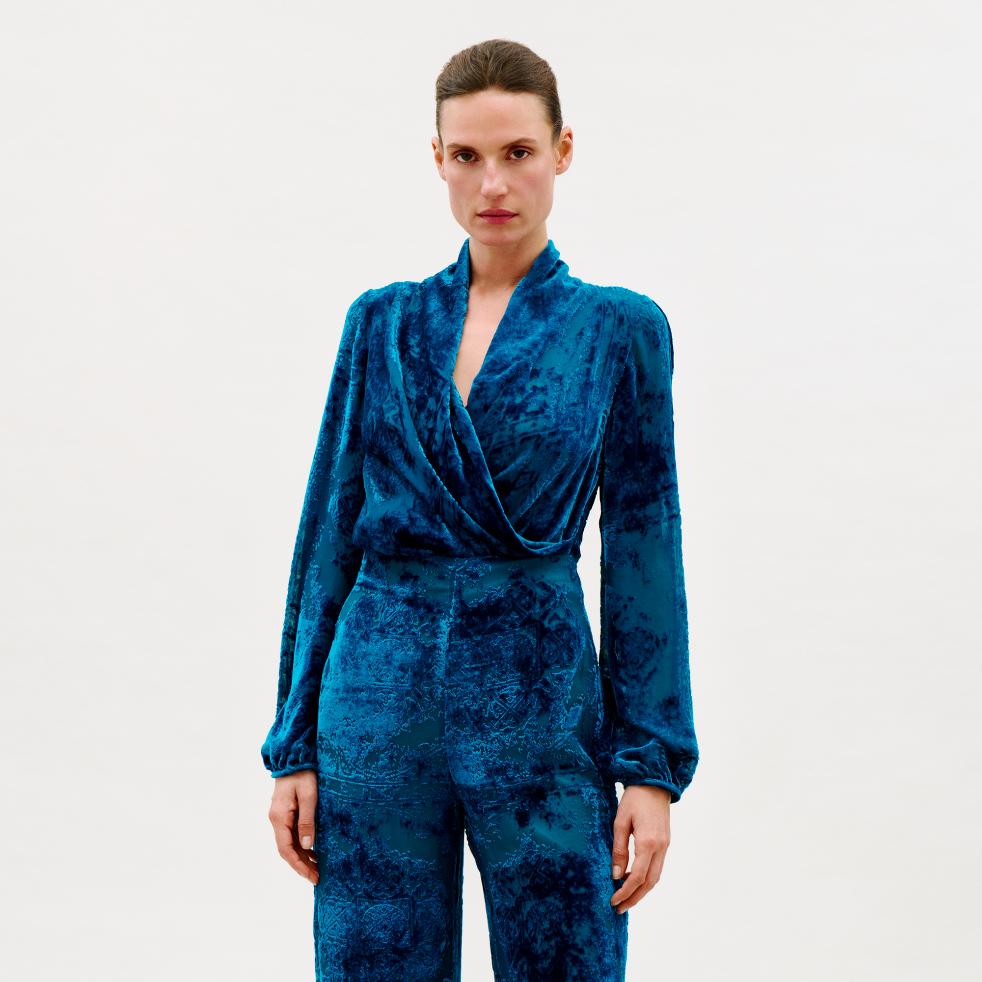 A woman stands against a plain light background, wearing the Tea Blouse in blue velvet with matching high-waisted pants. She looks directly at the camera with a neutral expression.