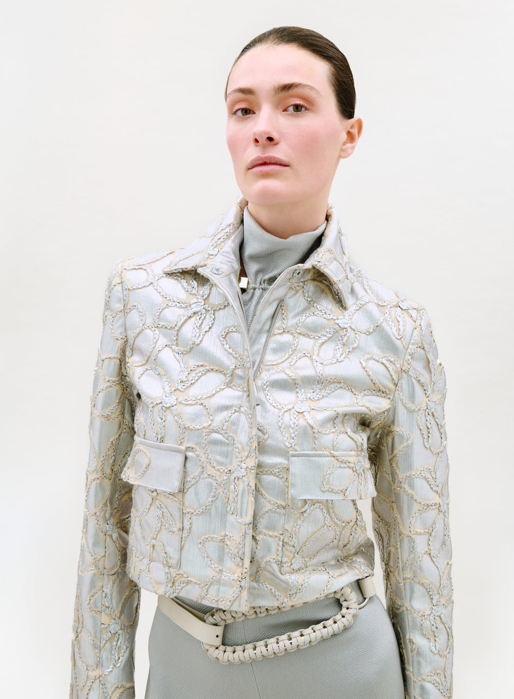 A person with slicked-back hair models the Perla Jacket—a light blue piece with gold embroidered floral patterns—over a turtleneck, paired with matching high-waisted pants and a braided belt, against a plain light background.