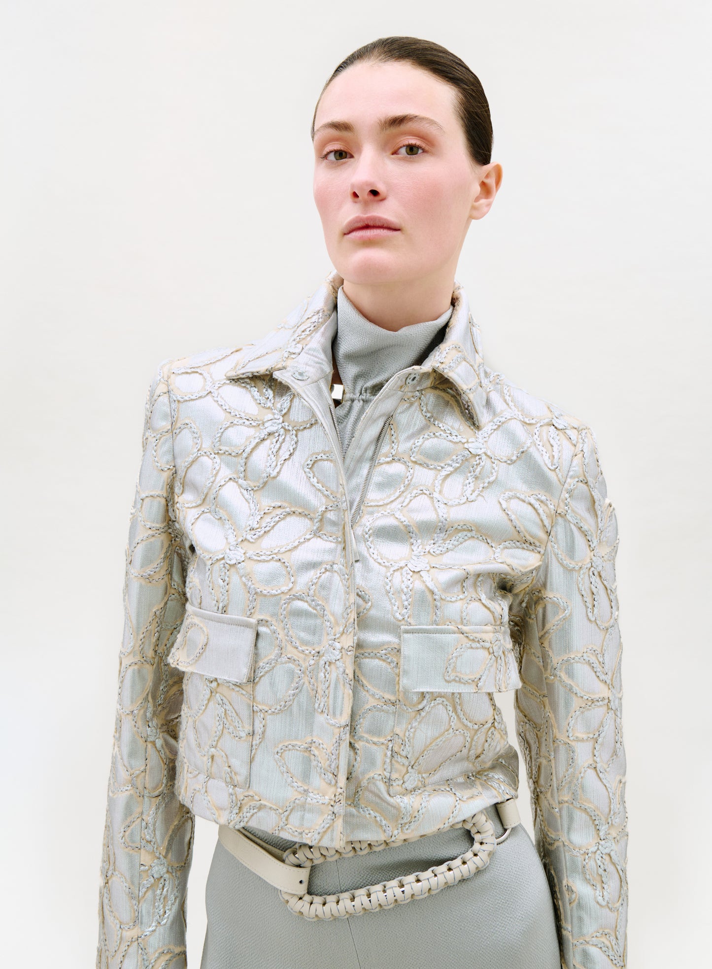 A person with slicked-back hair models the Perla Jacket—a light blue piece with gold embroidered floral patterns—over a turtleneck, paired with matching high-waisted pants and a braided belt, against a plain light background.