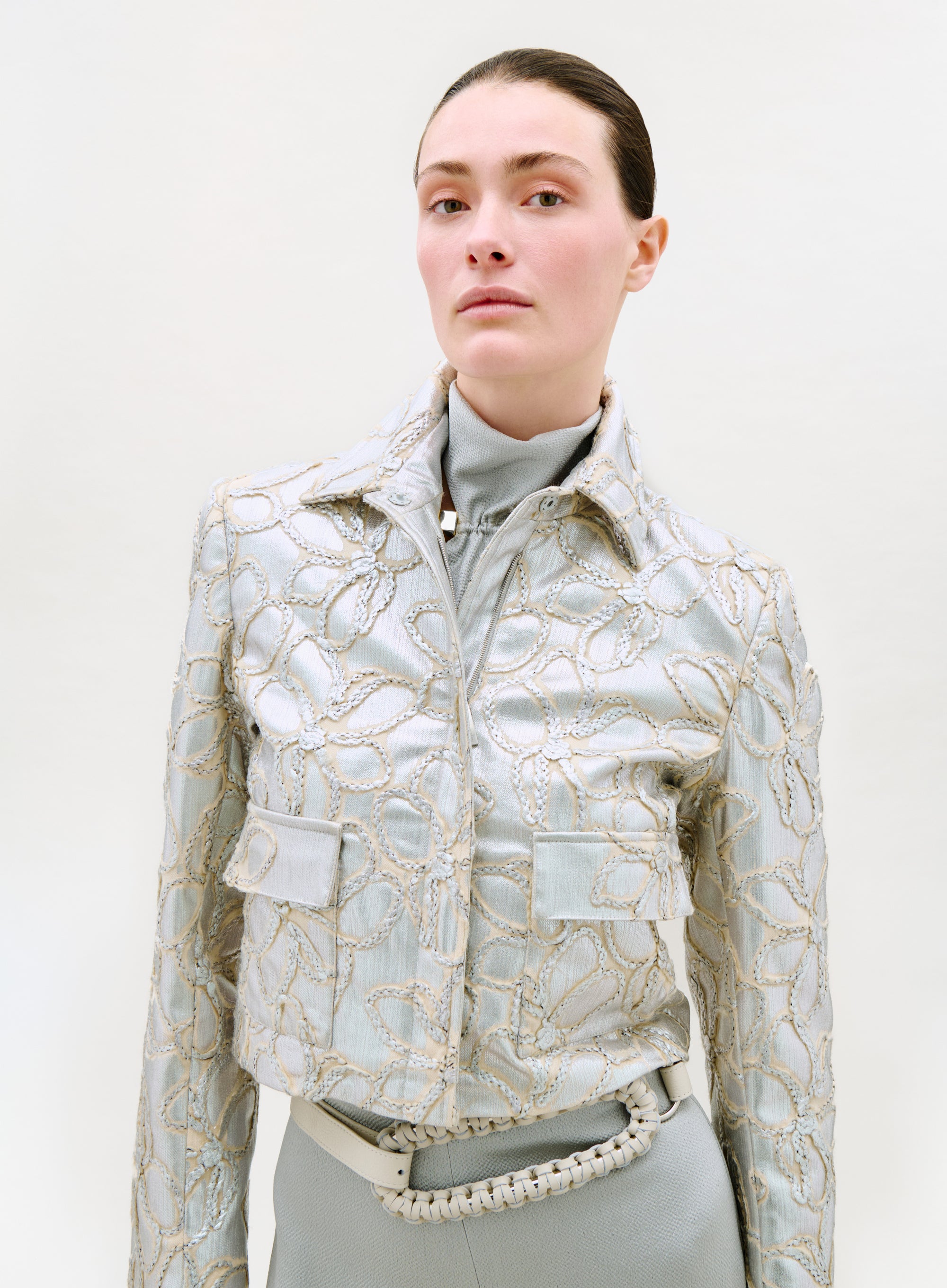 A person with slicked-back hair models the Perla Jacket—a light blue piece with gold embroidered floral patterns—over a turtleneck, paired with matching high-waisted pants and a braided belt, against a plain light background.