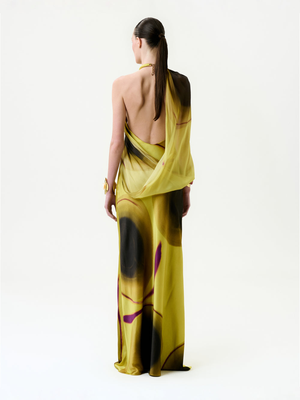 A person with long dark hair in a low ponytail models the Mari Luz Dress, a backless yellow silk maxi with black and purple abstract patterns. They are standing against a plain white background, facing away from the camera.