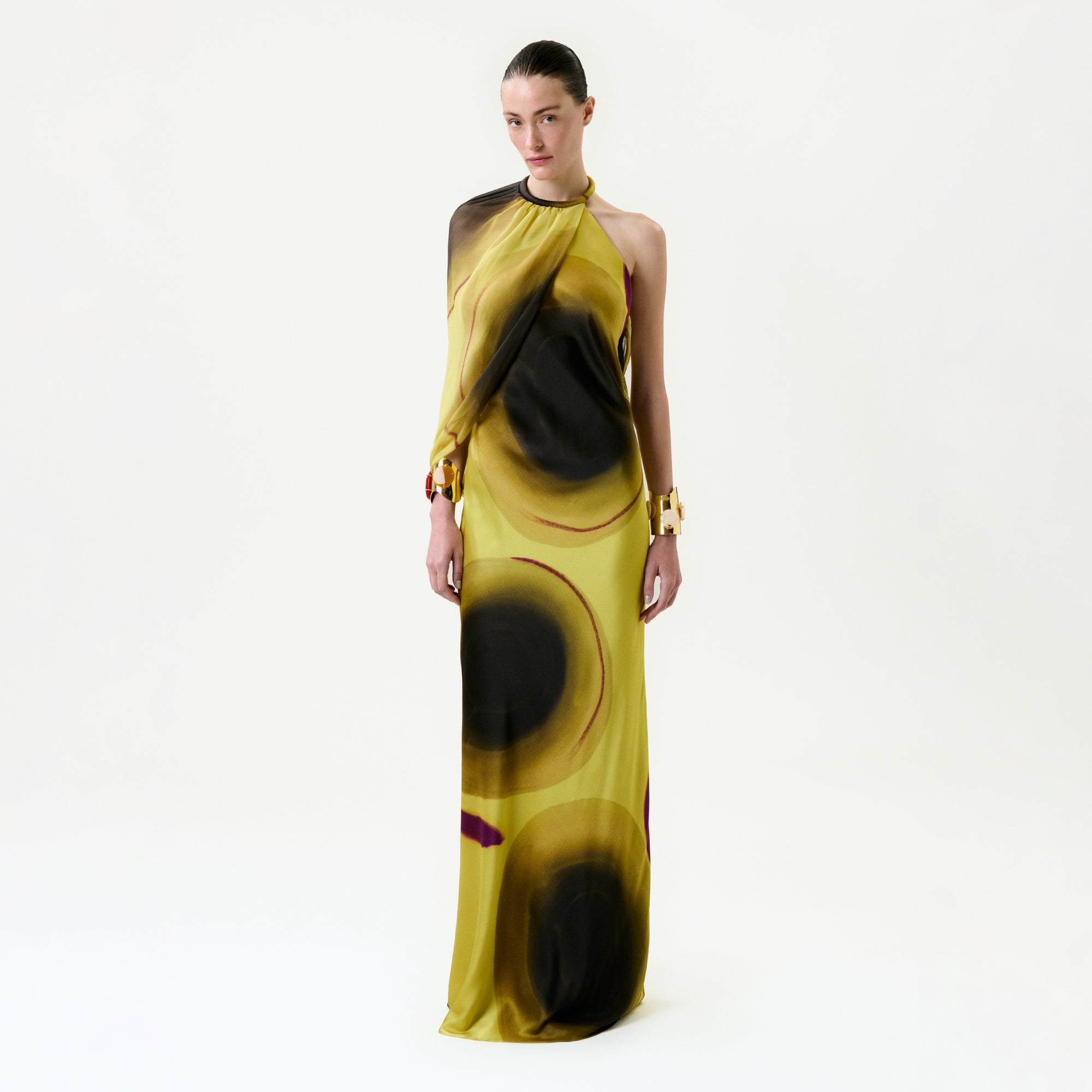A woman stands against a white background, wearing the Mari Luz Dress—a yellow, one-shoulder silk maxi dress with large black and brown abstract circular patterns and a halter neckline. She has slicked-back hair and gold cuff bracelets.