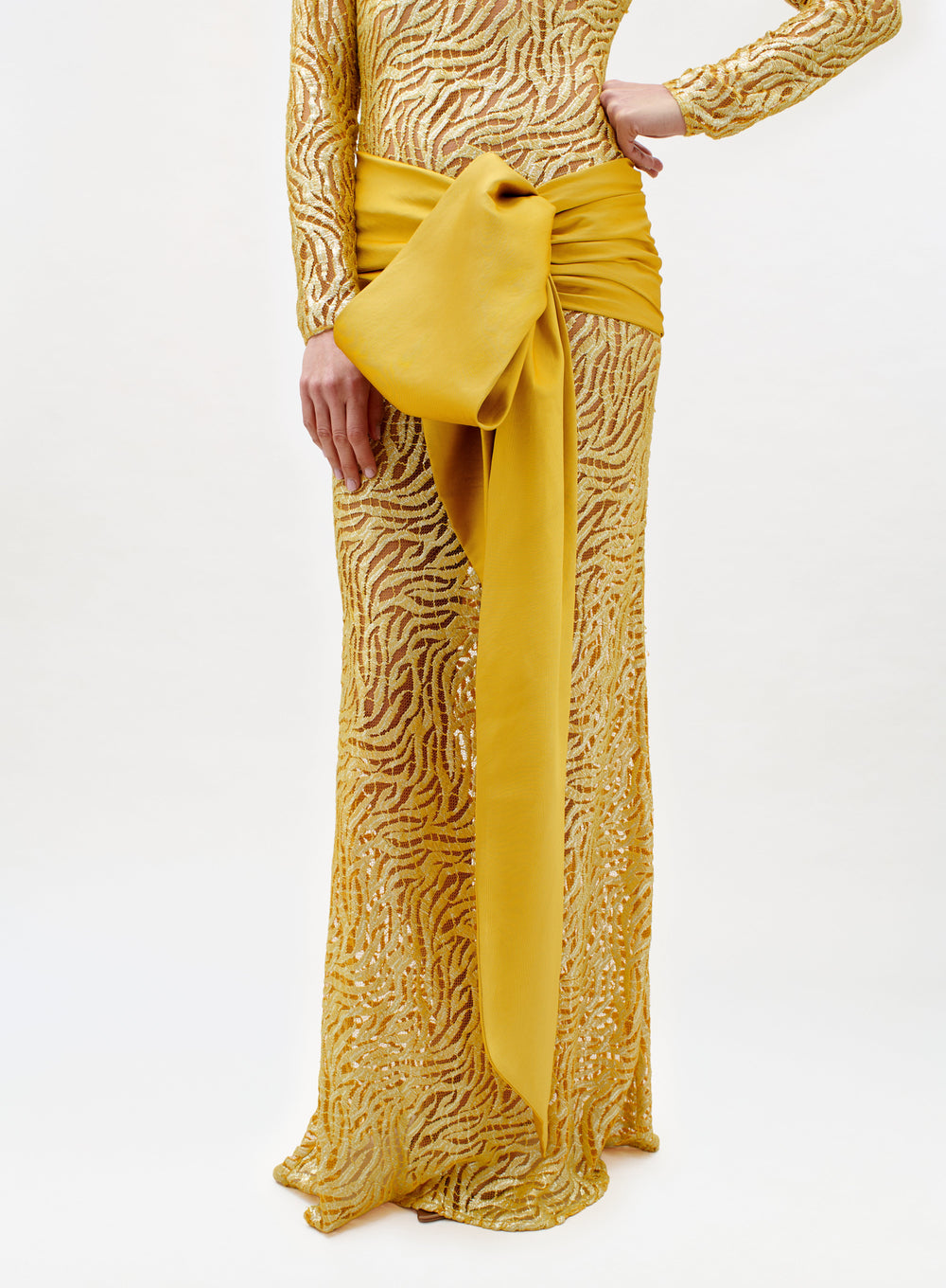 A person is wearing the Lia Dress—a long, gold sequined gown with a zebra stripe pattern and a bold yellow bow at the waist—while standing against a plain light background.