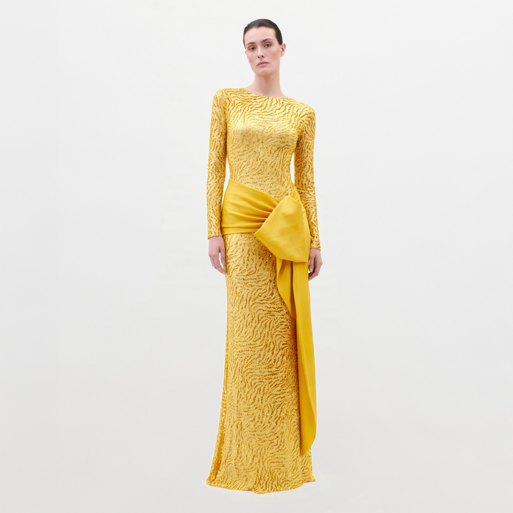 A woman stands against a plain light background, wearing the Lia Dress—a long-sleeved, floor-length yellow gown with a textured pattern and a bold asymmetrical satin bow at the waist.