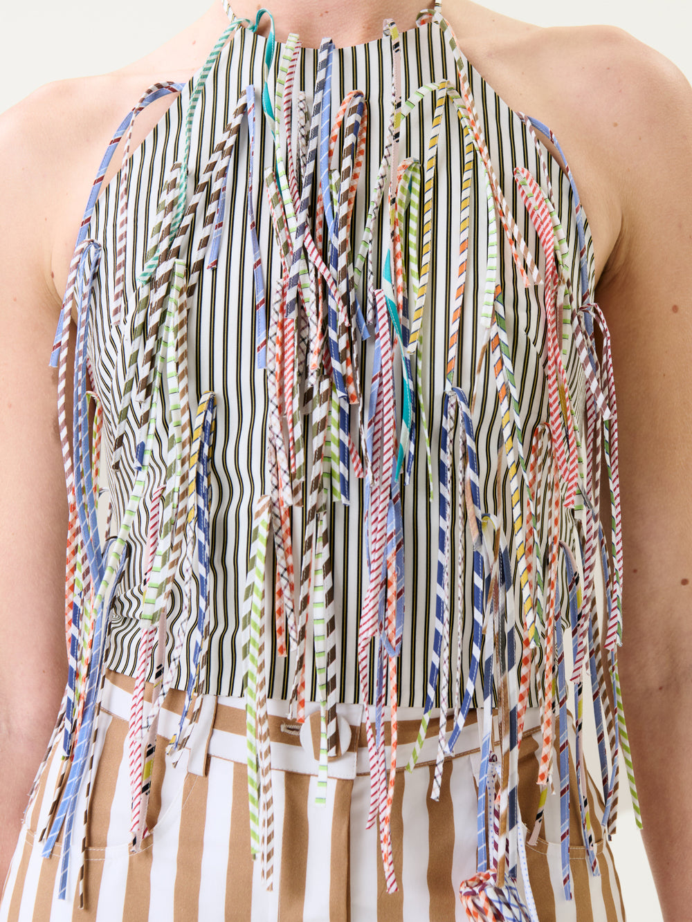 A person wearing the Lianne Top—a sleeveless top featuring vertical black and white stripes, colorful striped fringe hanging from the front, and tan and white striped fabric at the waist.