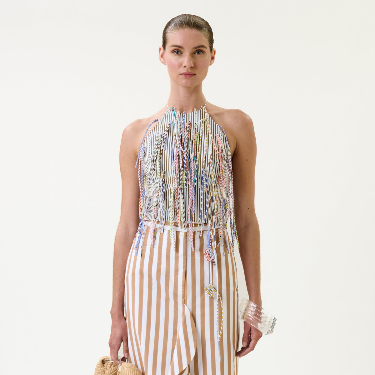 A woman wears the Lianne Top, a halter style with colorful fringe, paired with a beige-and-white vertical striped skirt. She holds a small handbag and a clear bracelet against a plain white background.