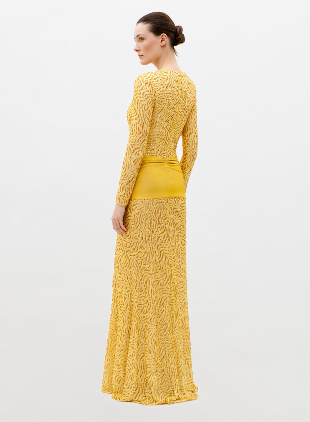 A woman with her hair in a neat bun wears the Lia Dress—a long-sleeved, floor-length yellow dress featuring a textured pattern and solid yellow waistband—standing against a plain light background.