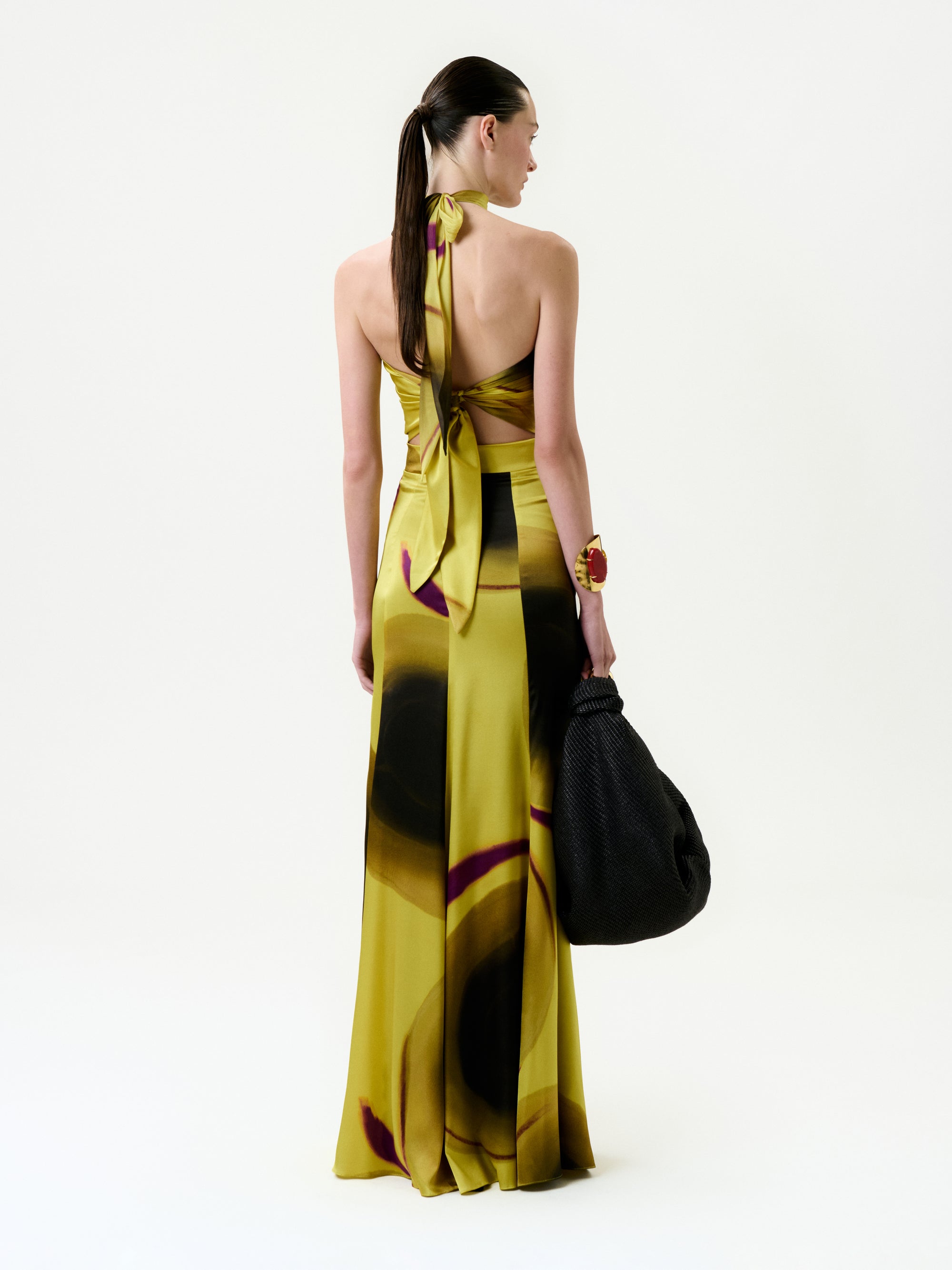 A woman with straight brown hair pulled back stands facing away, wearing the Marisol Skirt—a long, silky yellow skirt with abstract dark circular patterns. She holds a large black textured bag. Made in Portugal.