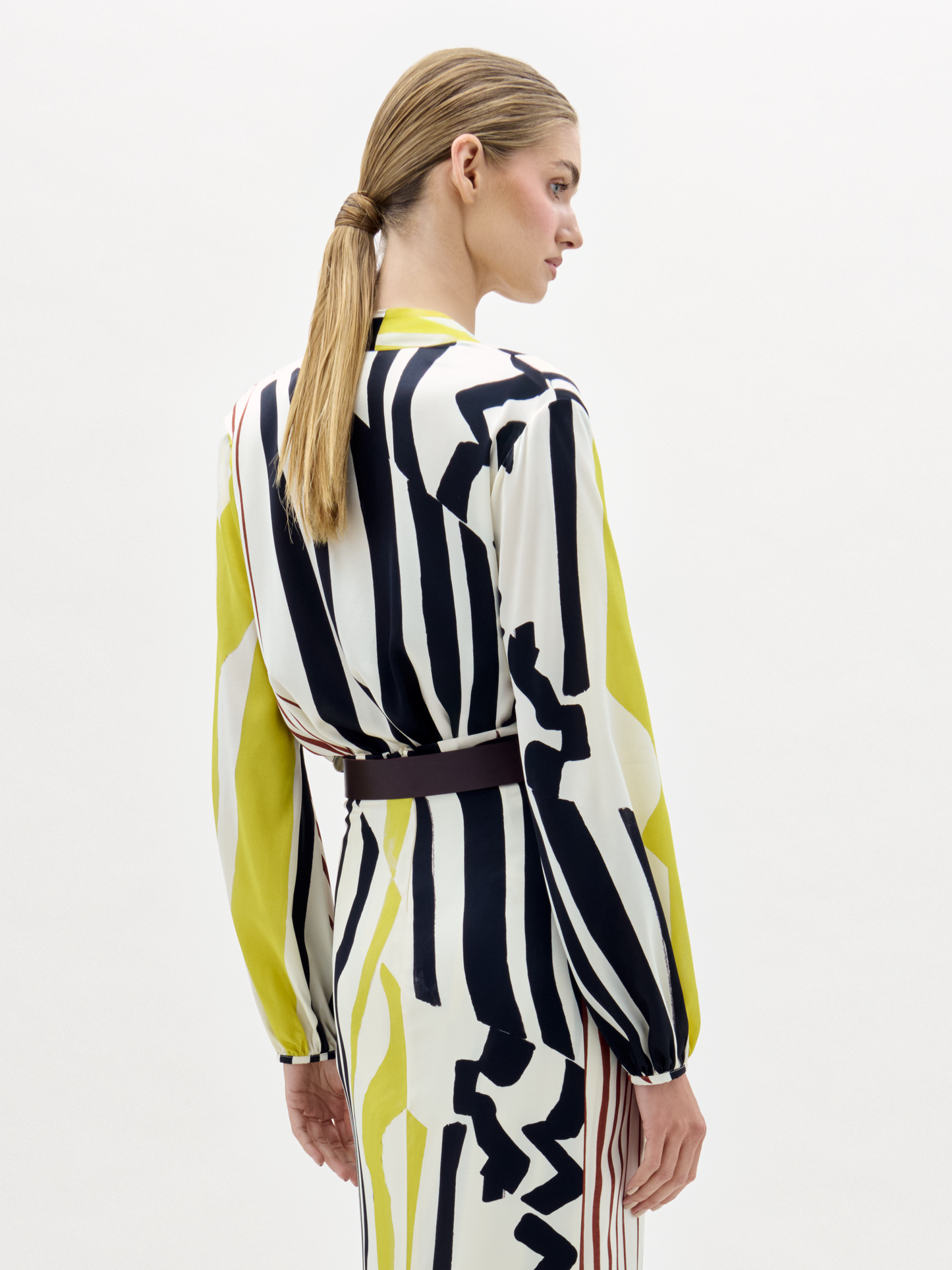 A woman poses against a plain background wearing the Honey Blouse, featuring long sleeves and a deep V-neck with black, white, yellow, and red stripes, styled with a wide belt.