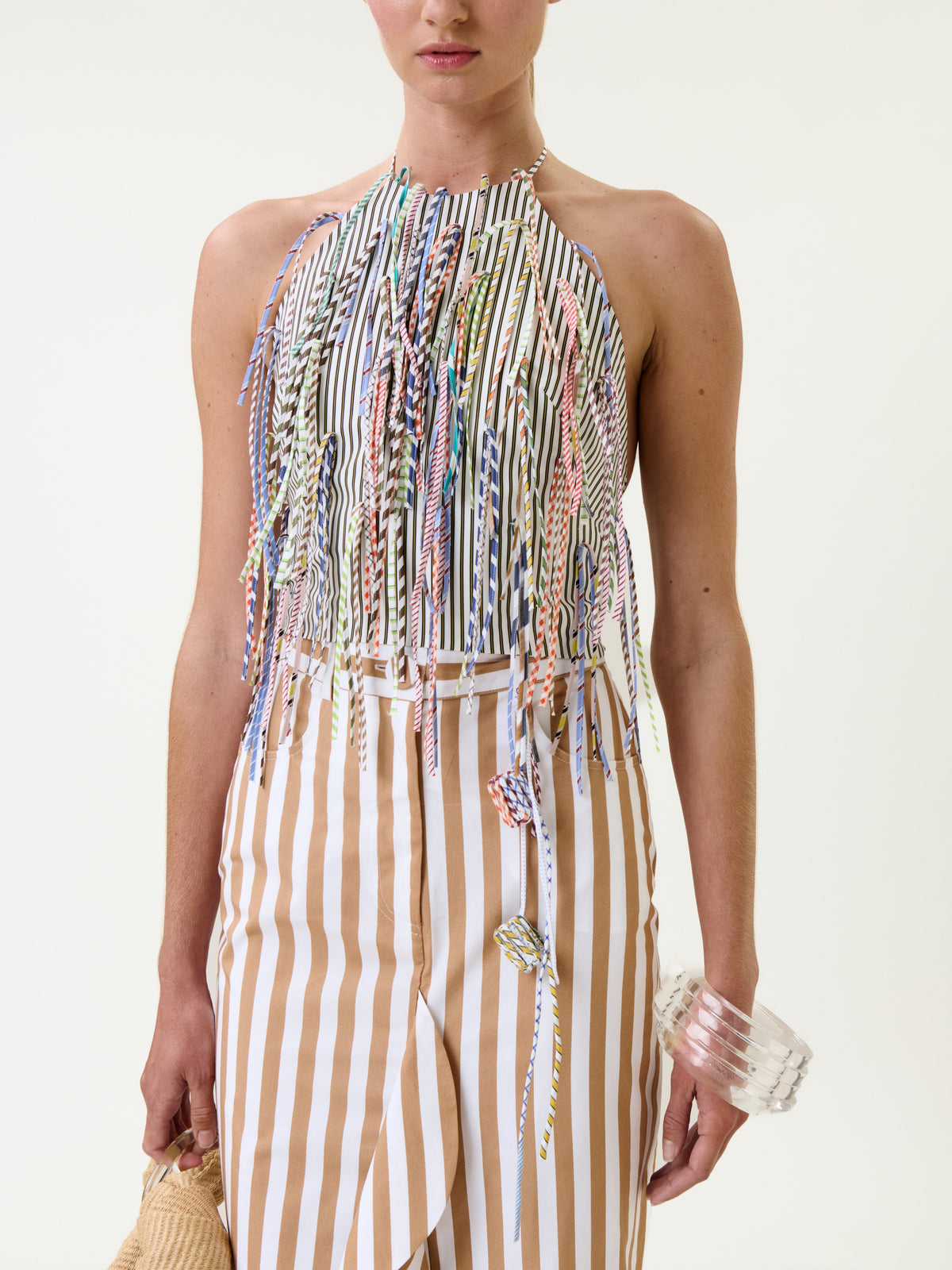 A woman wears a sleeveless top with colorful fringe and vertical stripes, paired with the Liuba Skirt—a tan-and-white striped skirt. She holds a clear bangle and a woven bag against a plain white background.