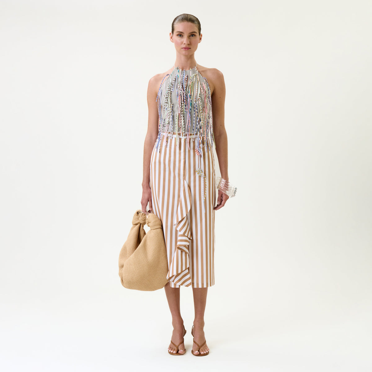 A woman stands against a plain background wearing a beaded halter top, the Liuba Skirt—a tan and white striped skirt with a ruffle—heeled sandals, and holding a large textured tan bag.