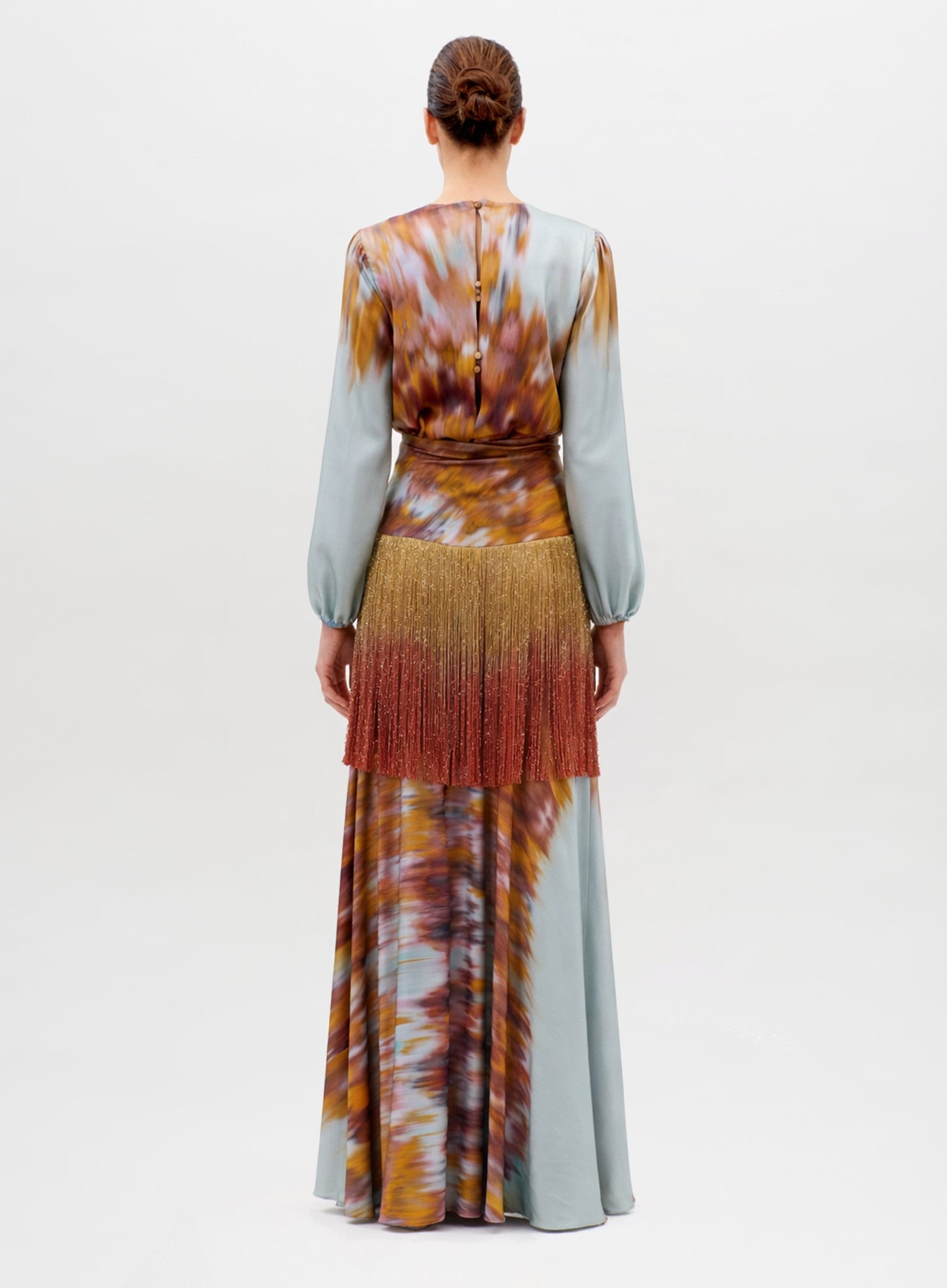 A woman stands facing away, wearing the Sutton Dress—a long, flowing piece with a colorful abstract pattern, light blue sleeves, gold and reddish fringe detail at the waist, and a long skirt.
