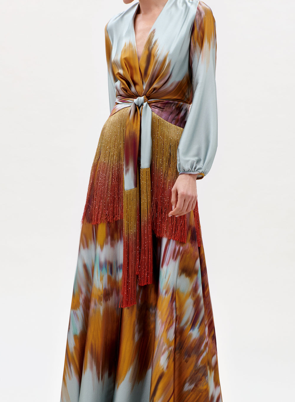 A model wears the Sutton Dress—a long, tie-dye dress with long sleeves, deep V-neck, tied waist, and red beaded fringe at the hips—standing against a plain light background.