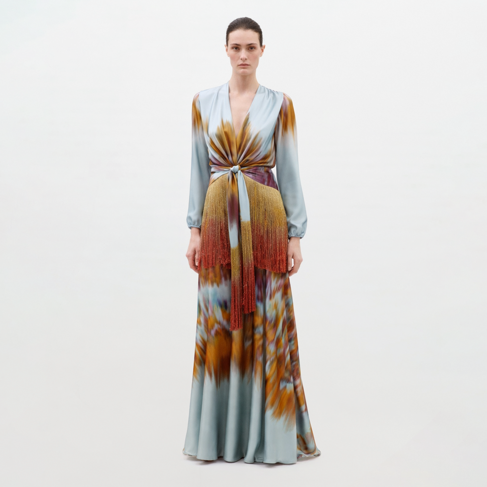 A woman poses against a plain background in the Sutton Dress, featuring blue, orange, and brown tie-dye patterns, long sleeves, and a knotted belt with fringe detail at the waist.