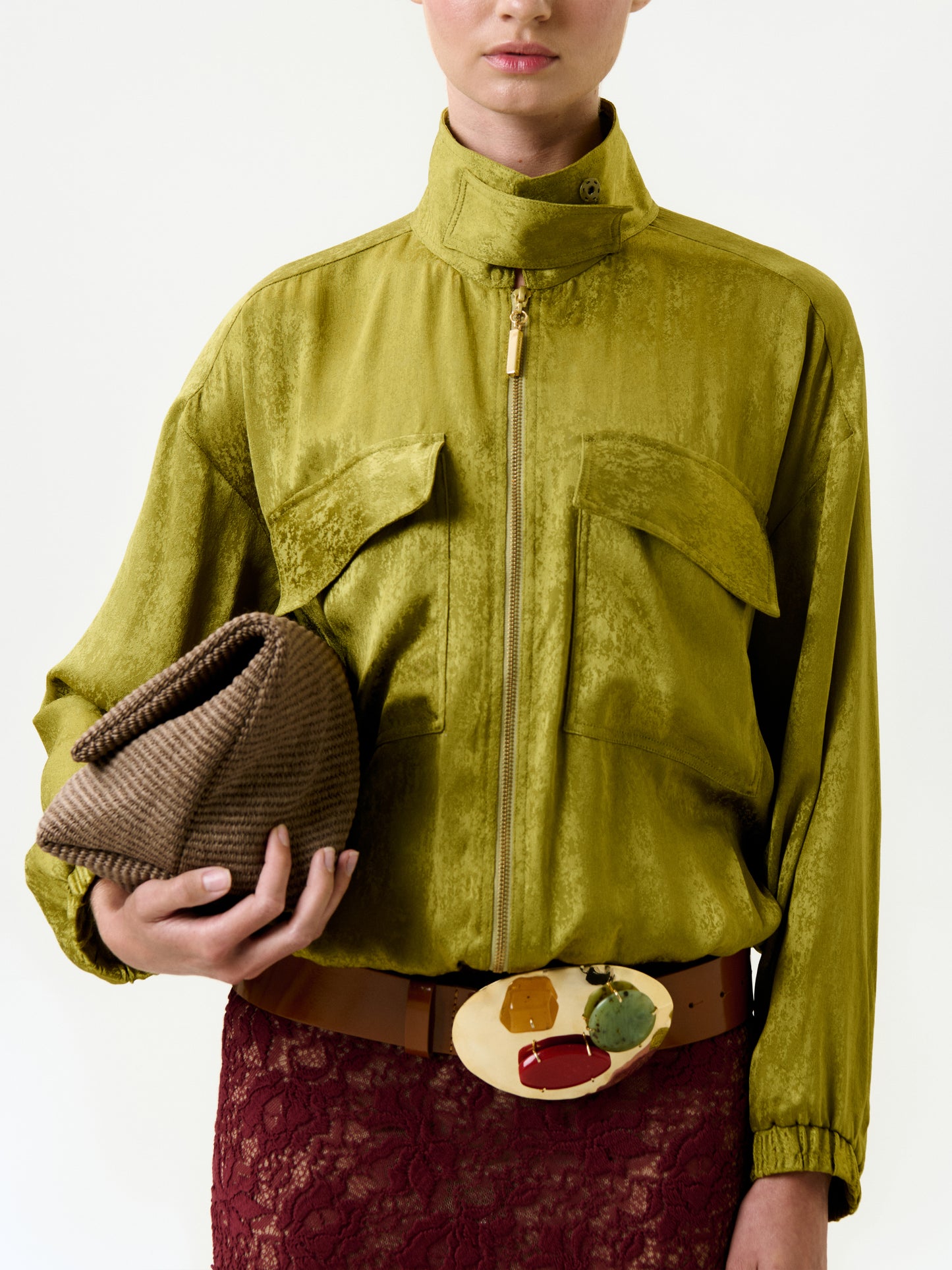 A person wears the Jhene Jacket in shiny olive green Italian fabric with large front pockets, paired with a brown and gold statement belt, red lace skirt, and a textured brown 100% viscose clutch bag against a plain background.