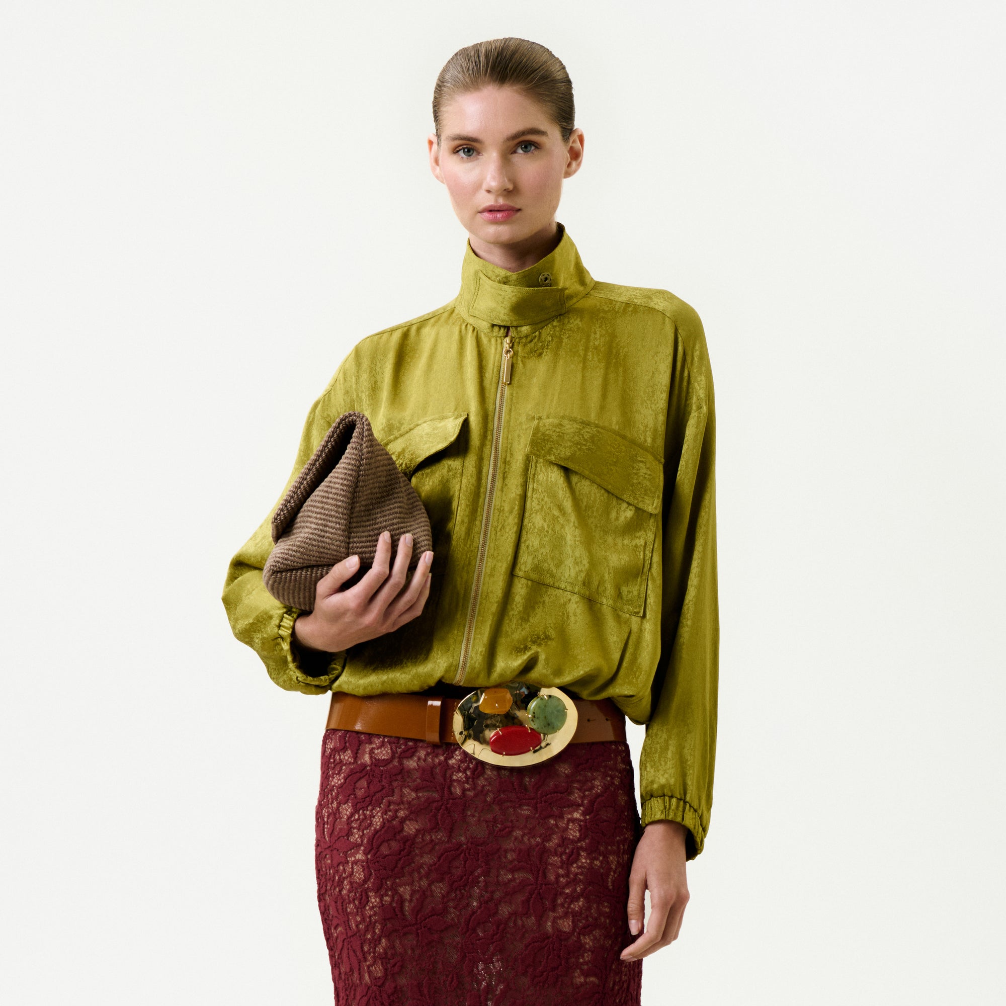 A woman in the Jhene Jacket—Italian fabric, 100% viscose, shiny olive-green, large pockets—pairs it with a brown lace skirt, wide belt with round buckle, and textured clutch. Made in Colombia.