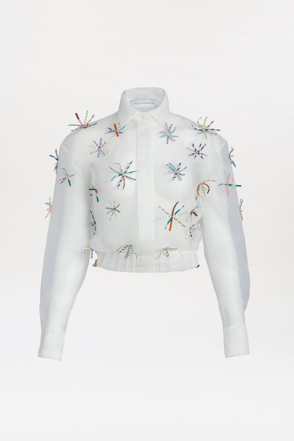 The Katniss Jacket is a white, long-sleeve collared piece featuring colorful embroidered starburst patterns and small beads scattered across the slightly sheer fabric. It has a button-down front for easy wear.