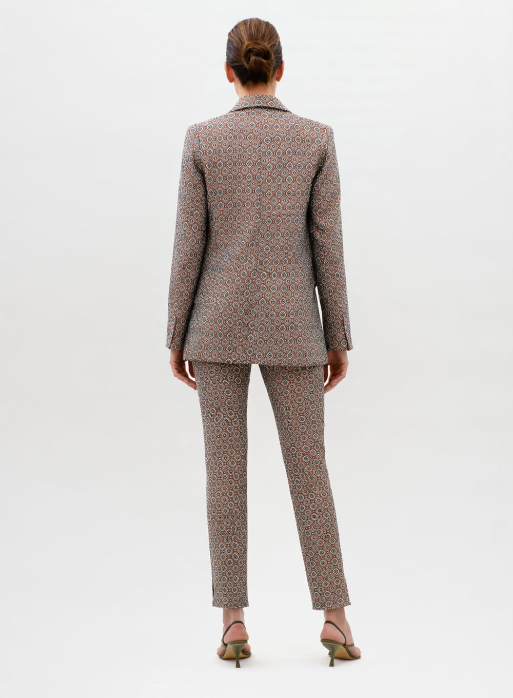 A woman stands facing away, wearing the Cuneo Jacket as part of a patterned suit with matching straight-leg pants. Her hair is tied back, and she pairs the outfit with heeled sandals against a plain, light background.