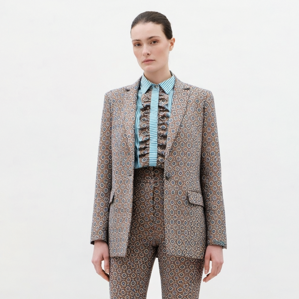 A woman with straight brown hair pulled back and a neutral expression stands against a plain background wearing the Cuneo Jacket, paired with matching patterned pants and a blue striped ruffled shirt.