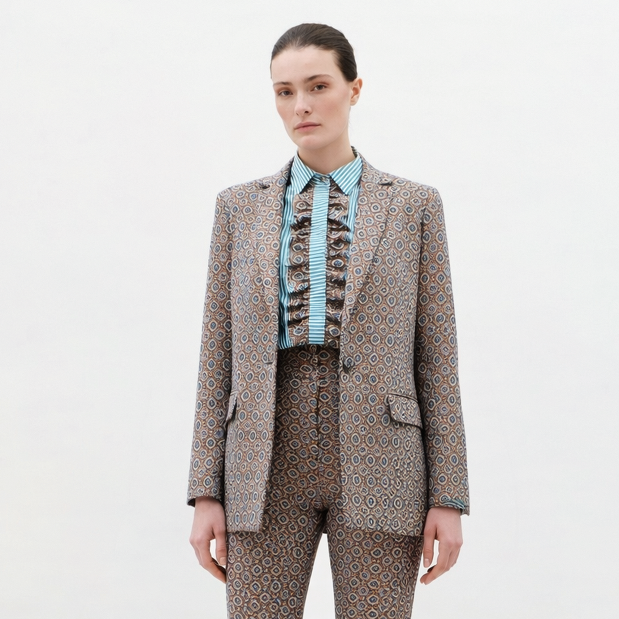 A woman with straight brown hair pulled back and a neutral expression stands against a plain background wearing the Cuneo Jacket, paired with matching patterned pants and a blue striped ruffled shirt.