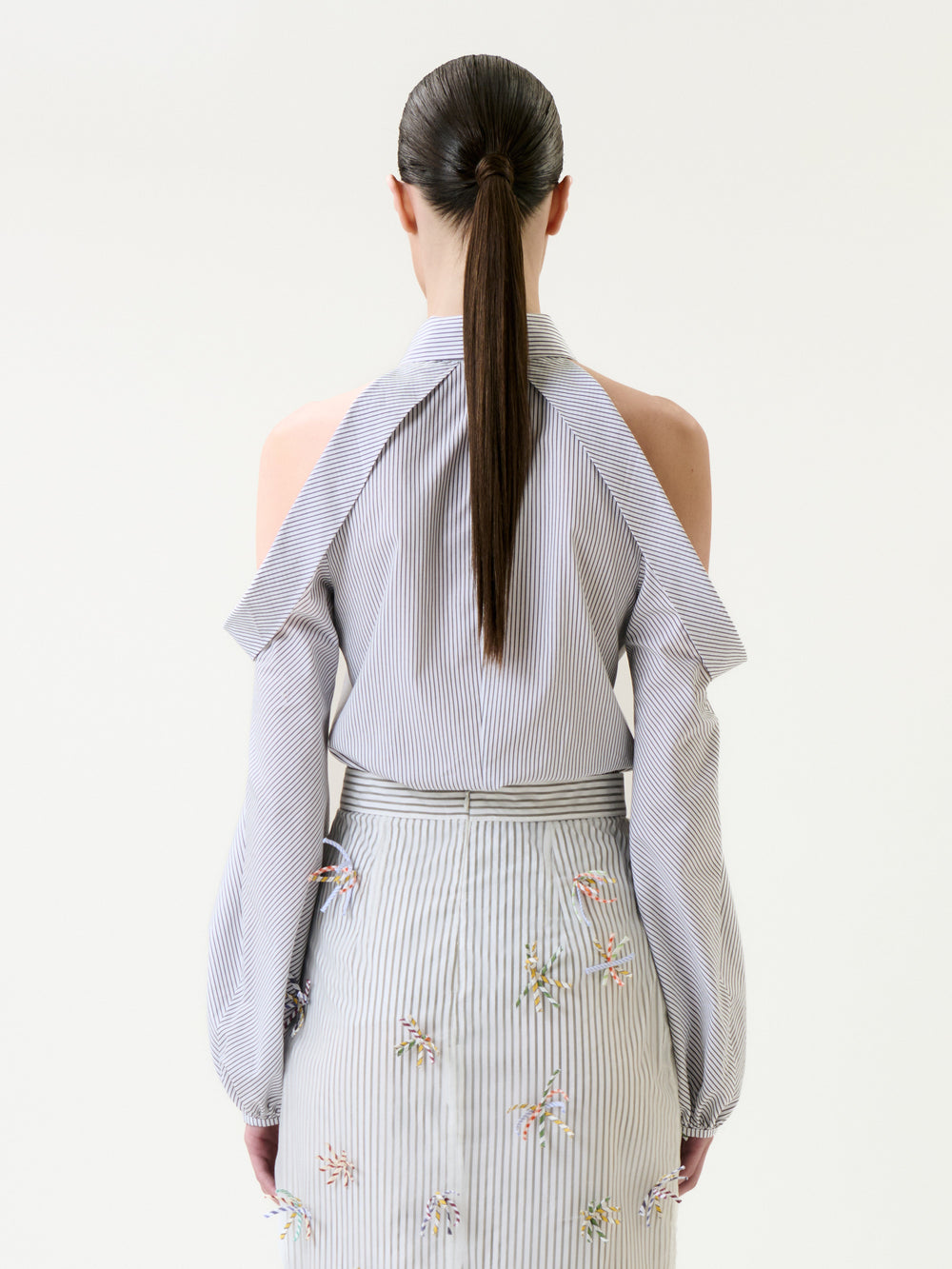 A woman with a long ponytail is shown from behind, wearing the Nurith Shirt—a striped blouse with cold-shoulder sleeves—paired with a matching skirt decorated with small colorful embroidered patterns.