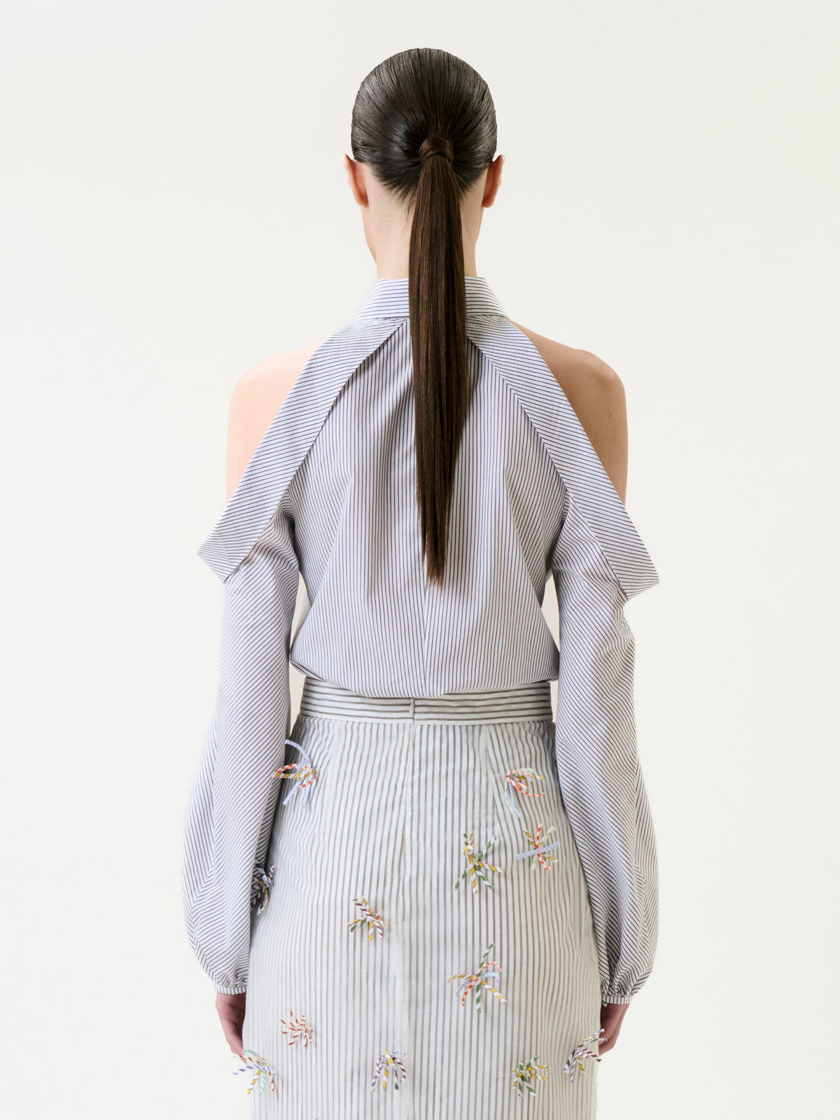 A woman with a long ponytail is shown from behind, wearing the Nurith Shirt—a striped blouse with cold-shoulder sleeves—paired with a matching skirt decorated with small colorful embroidered patterns.
