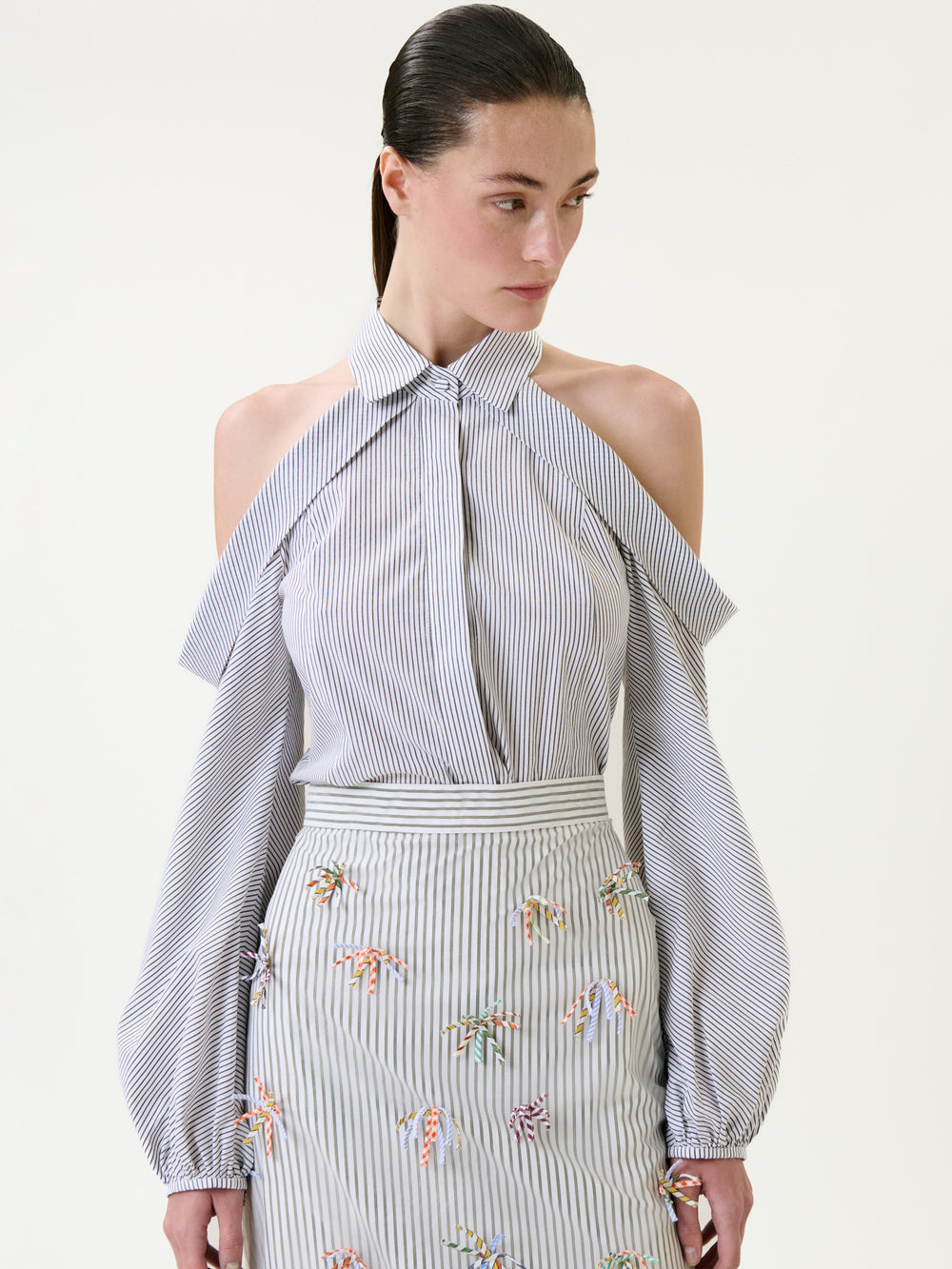 Wearing the Nurith Shirt—a blue-and-white striped blouse with cold-shoulder sleeves—paired with a matching skirt adorned with colorful embroidered details, the woman stands against a light background.