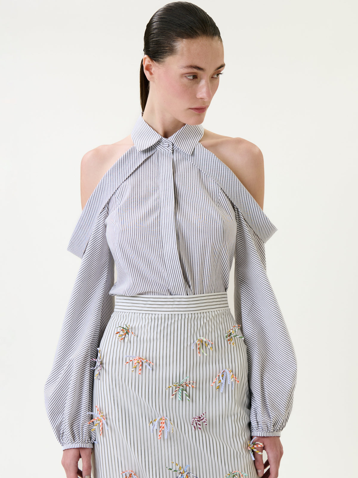 Wearing the Nurith Shirt—a blue-and-white striped blouse with cold-shoulder sleeves—paired with a matching skirt adorned with colorful embroidered details, the woman stands against a light background.