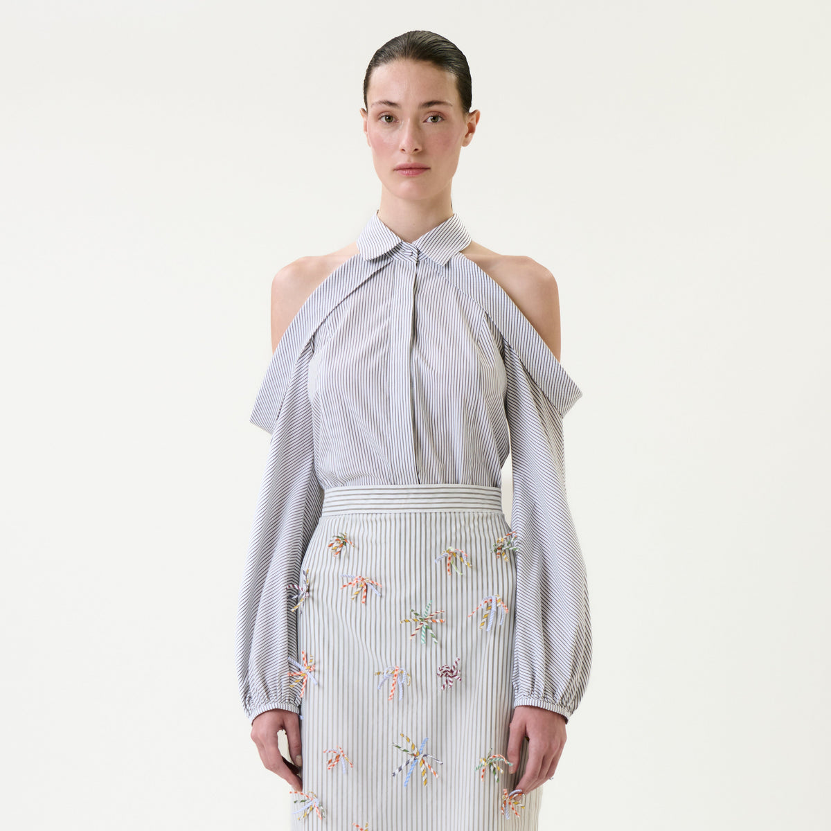 A woman wears the Nurith Shirt, a light blue cold-shoulder blouse with draped sleeves, paired with a matching high-waisted skirt adorned with small, colorful embroidered flowers, standing against a plain white background.