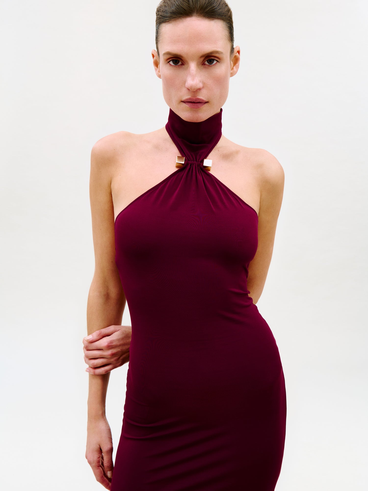 A model wearing a sleeveless, high-neck halter dress in deep burgundy, photographed against a clean white studio background.