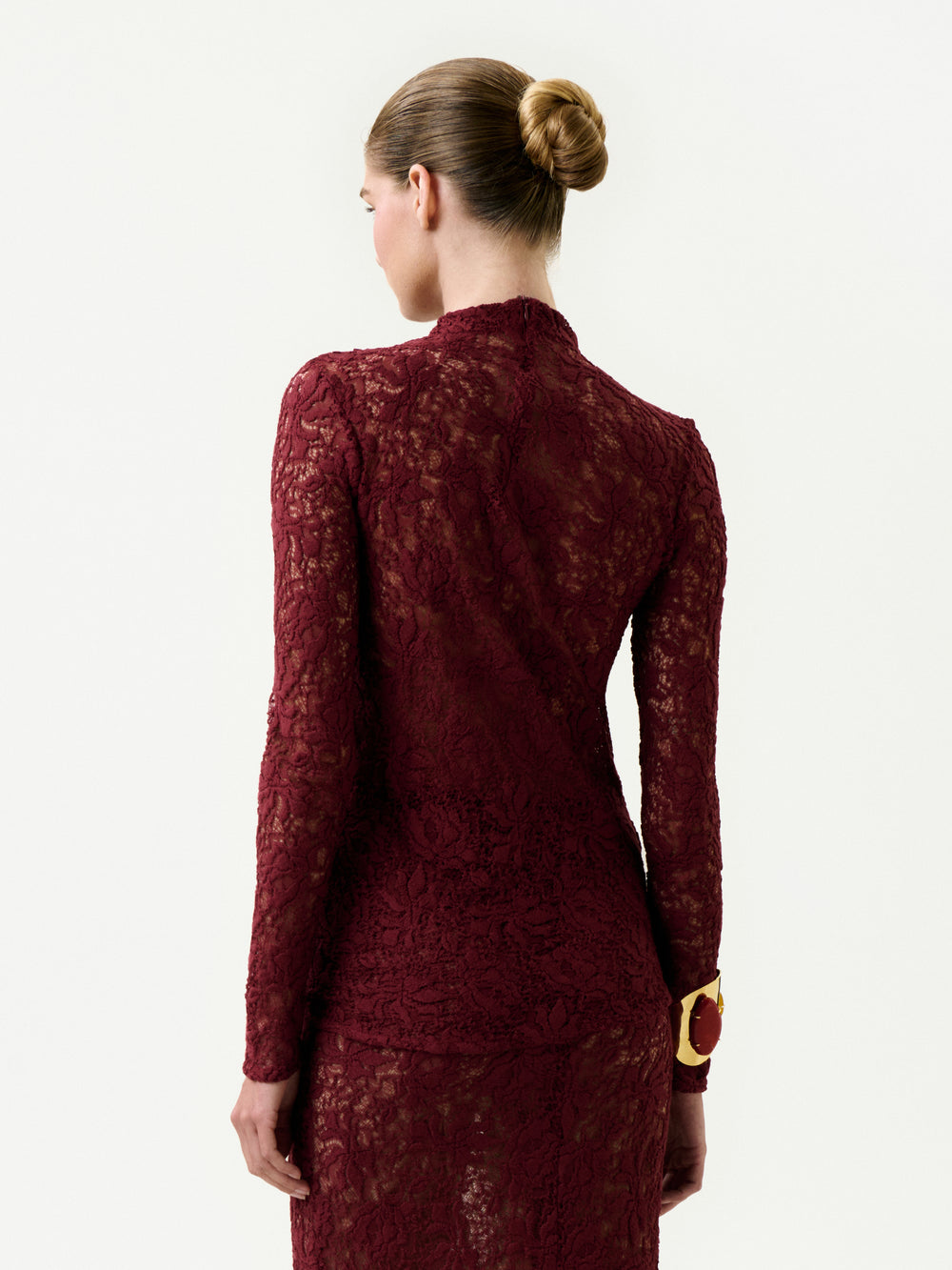 A woman with her hair in a neat bun, wearing the Lisibel Blouse—an elegant, long-sleeved burgundy lace top made from Italian fabric—stands facing away from the camera against a plain white background.