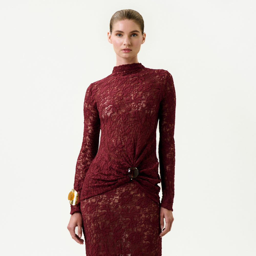 A woman wearing the Lisibel Blouse—burgundy, long-sleeved, high-neck, crafted from fine Italian lace with a brooch detail at the waist—stands against a light background. Her hair is pulled back and she wears a large bracelet on her right wrist.