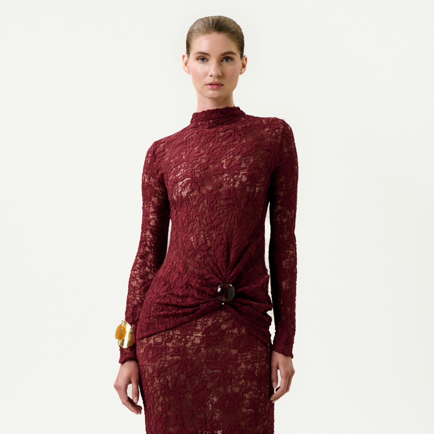 A woman wearing the Lisibel Blouse—burgundy, long-sleeved, high-neck, crafted from fine Italian lace with a brooch detail at the waist—stands against a light background. Her hair is pulled back and she wears a large bracelet on her right wrist.