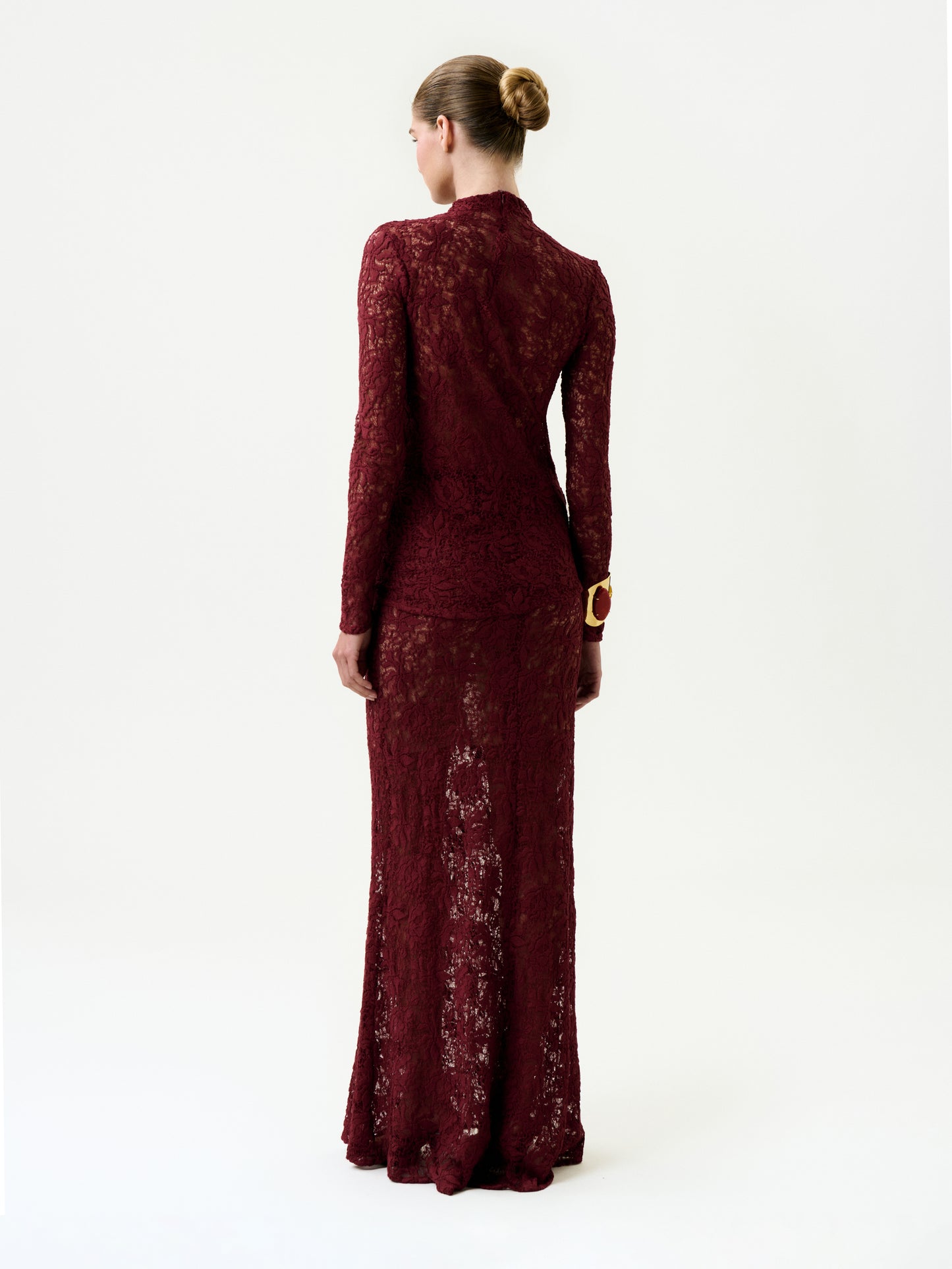 A woman with her hair in a bun stands facing away, wearing the floor-length, long-sleeved Malek Skirt in luxurious Italian burgundy lace with a fitted silhouette, set against a plain white background.