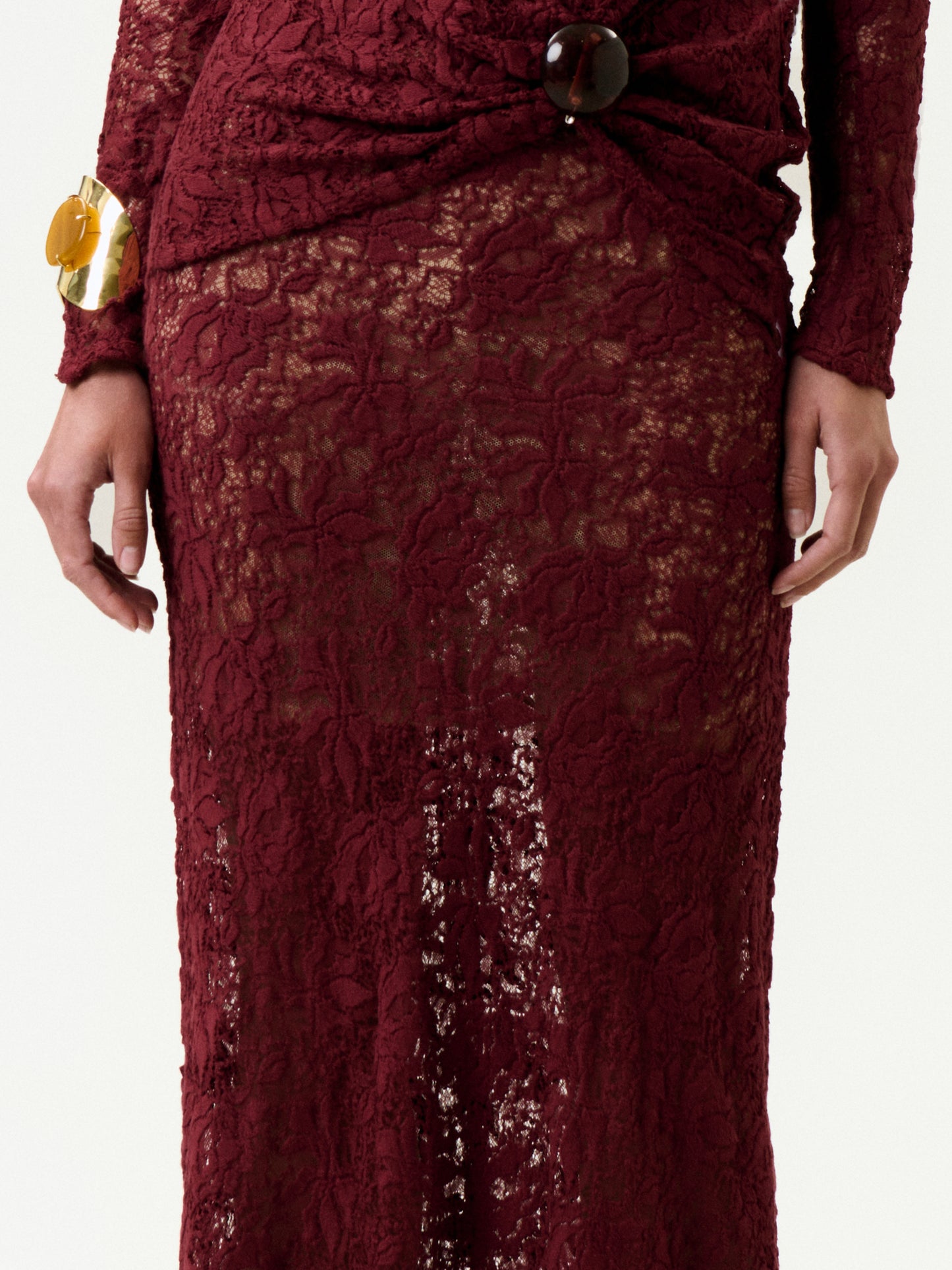 A person wearing the Malek Skirt, a long burgundy lace piece with intricate patterns made in Colombia, featuring a gathered waist with a large round button and paired with a chunky gold bracelet on their left wrist.