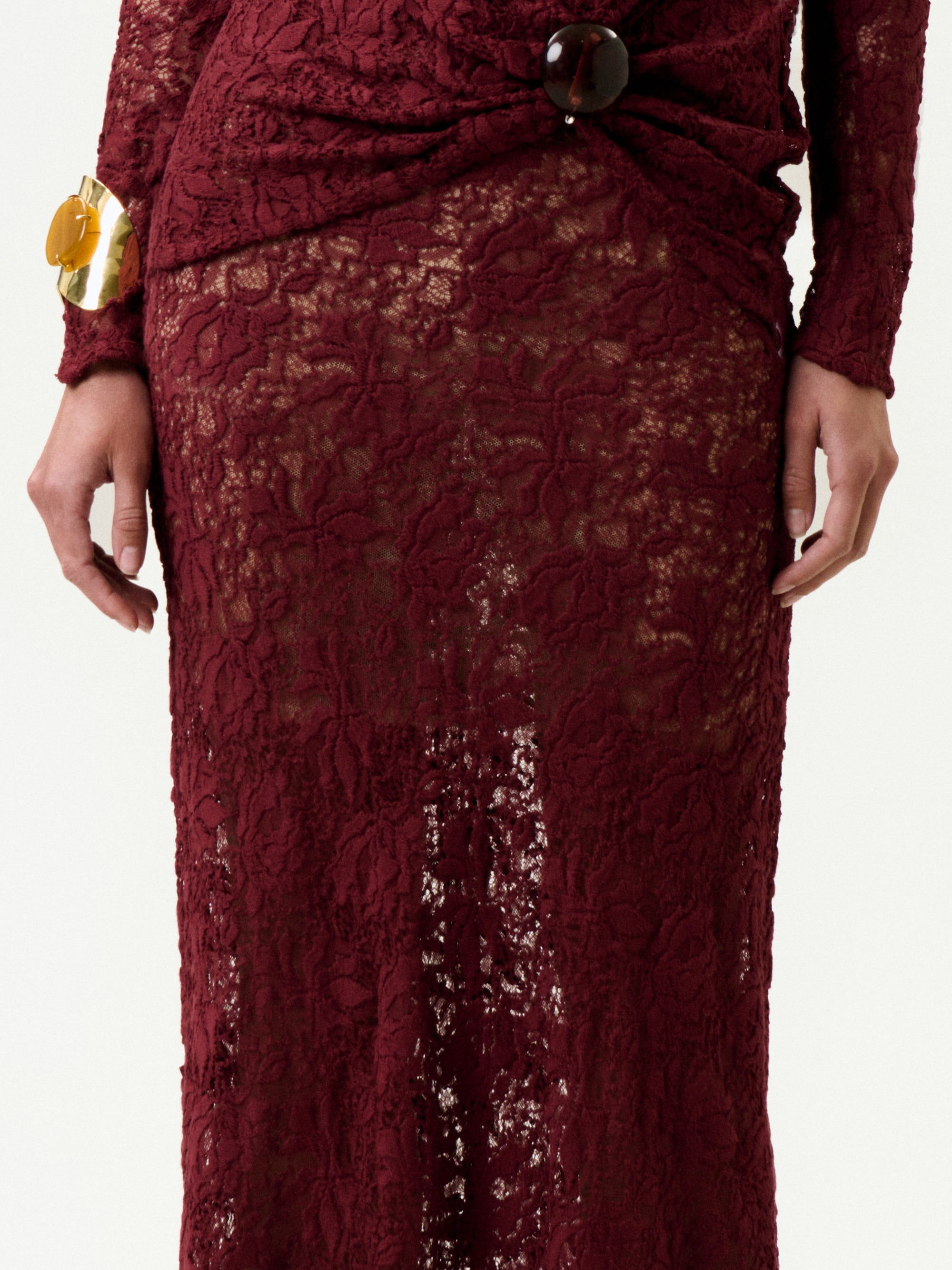 A person wearing the Malek Skirt, a long burgundy lace piece with intricate patterns made in Colombia, featuring a gathered waist with a large round button and paired with a chunky gold bracelet on their left wrist.