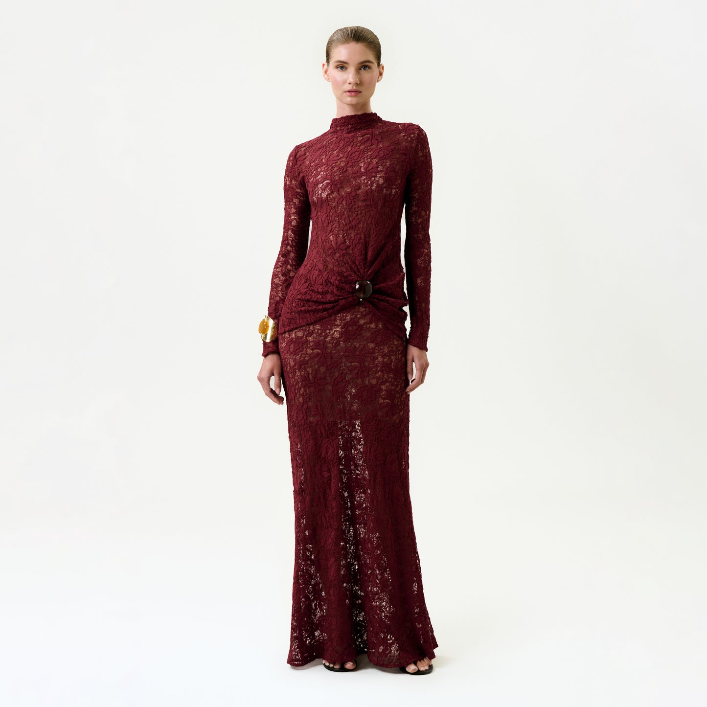 A woman stands against a plain background wearing the Malek Skirt, a long burgundy lace skirt crafted from luxurious Italian fabric, paired with a gold bracelet and black open-toe heels.