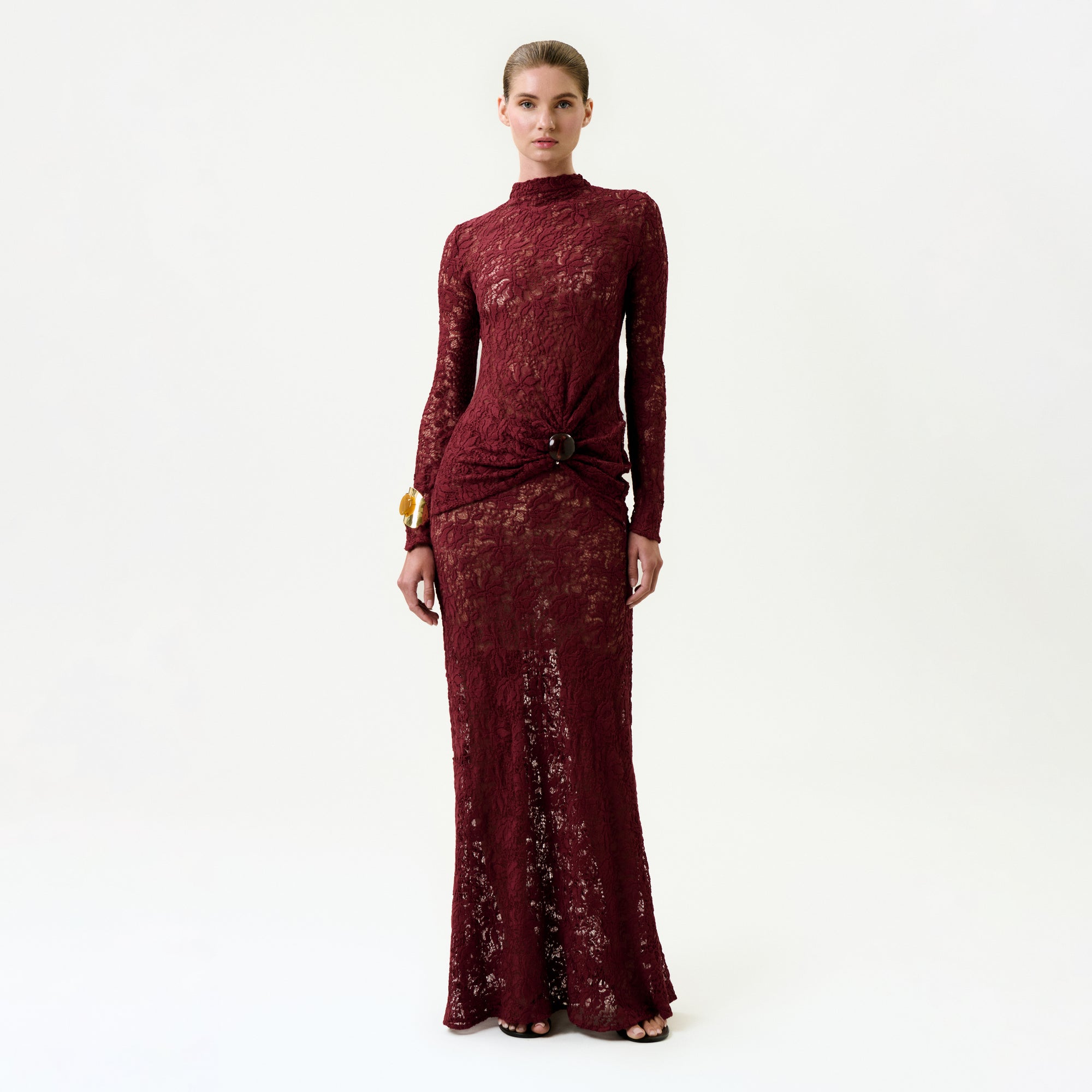 A woman stands against a plain background wearing the Malek Skirt, a long burgundy lace skirt crafted from luxurious Italian fabric, paired with a gold bracelet and black open-toe heels.