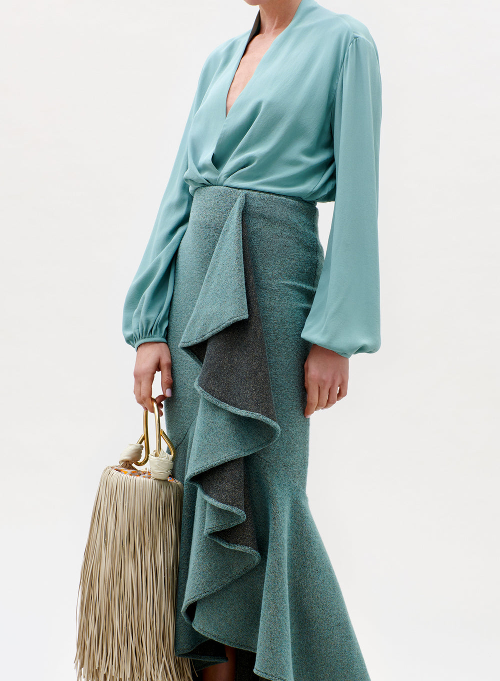 A woman models the Delfina Blouse in light teal, paired with a high-waisted ruffle skirt and holding a beige fringed handbag. Her face is not visible in the image.