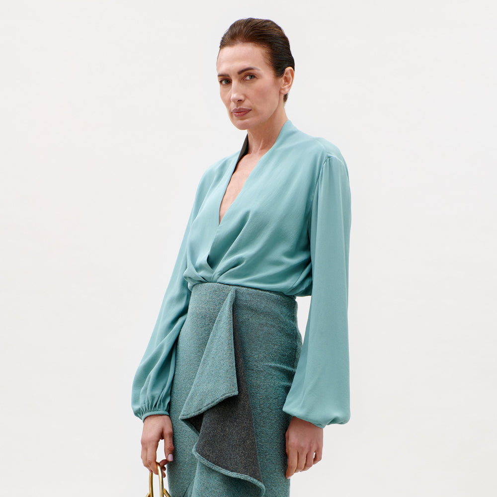 A woman with dark hair pulled back wears the Delfina Blouse—a light teal top with balloon sleeves—paired with a textured, asymmetrical teal skirt, standing against a plain light background.