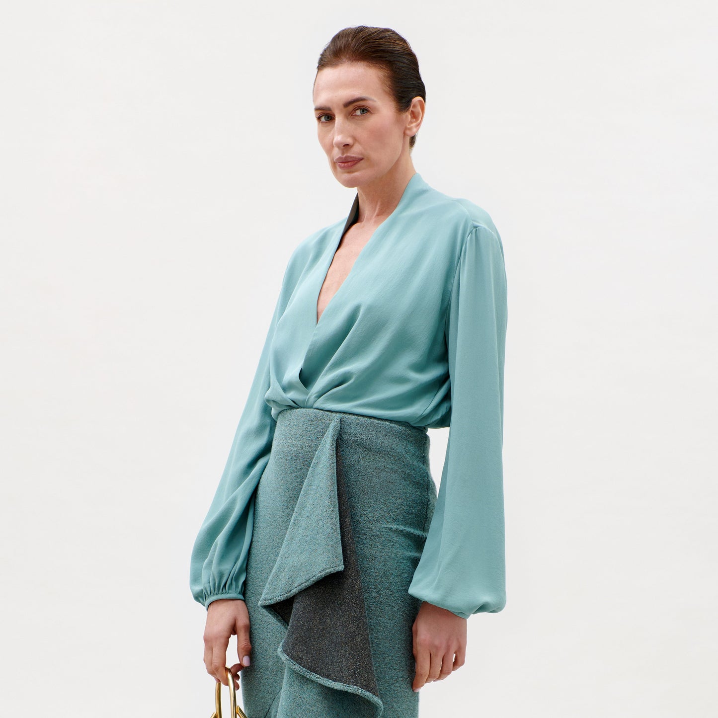 A woman with dark hair pulled back wears the Delfina Blouse—a light teal top with balloon sleeves—paired with a textured, asymmetrical teal skirt, standing against a plain light background.