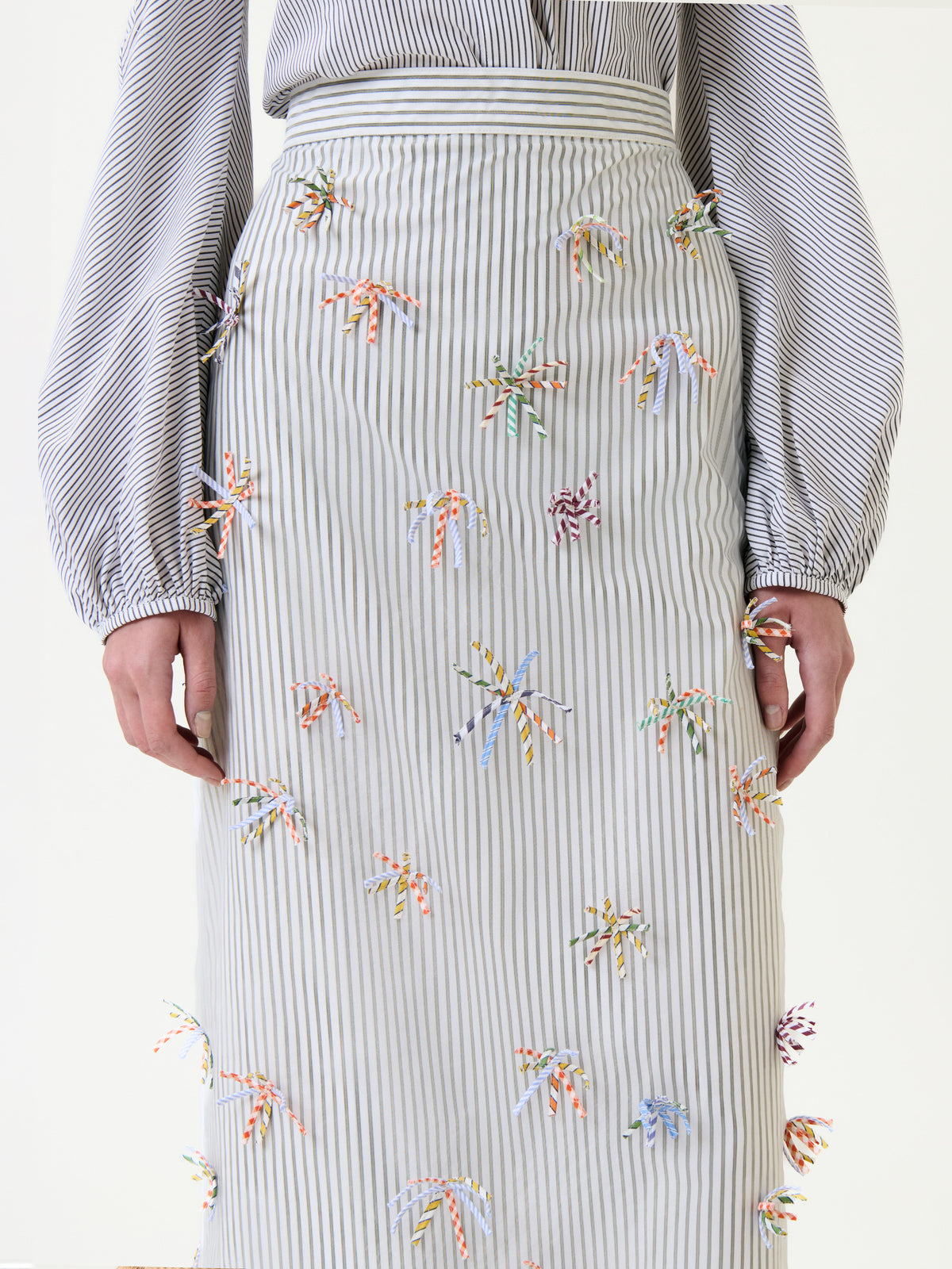 The photo shows a person wearing the Uma Skirt, styled with a striped shirt. The skirt is adorned with small, colorful, knotted ribbons, and the cropped image highlights the textured details of the outfit from shoulders to knees.
