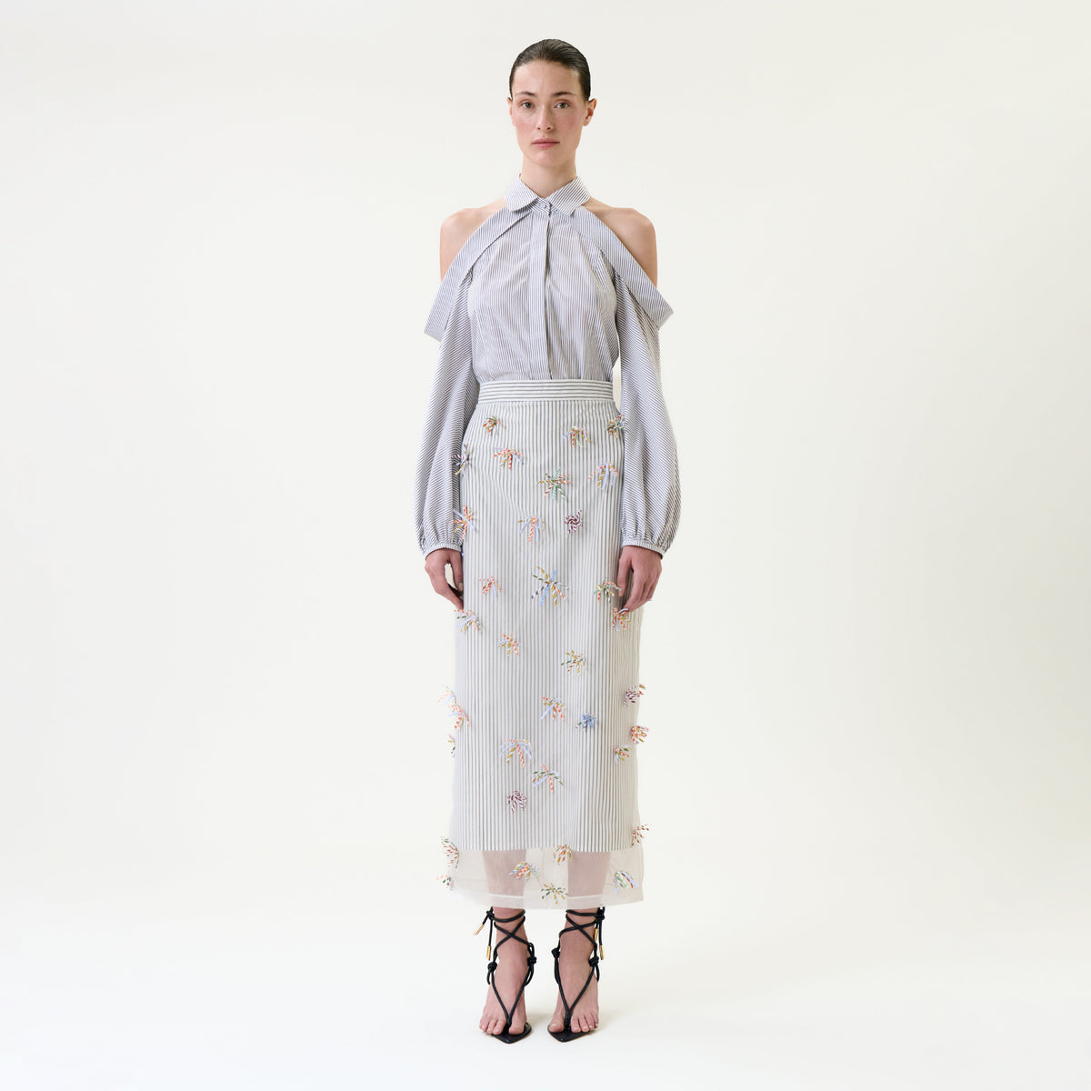 A woman stands against a plain background wearing a light blue, cold-shoulder blouse and the Uma Skirt—a sheer embroidered skirt with floral patterns—paired with black strappy sandals.