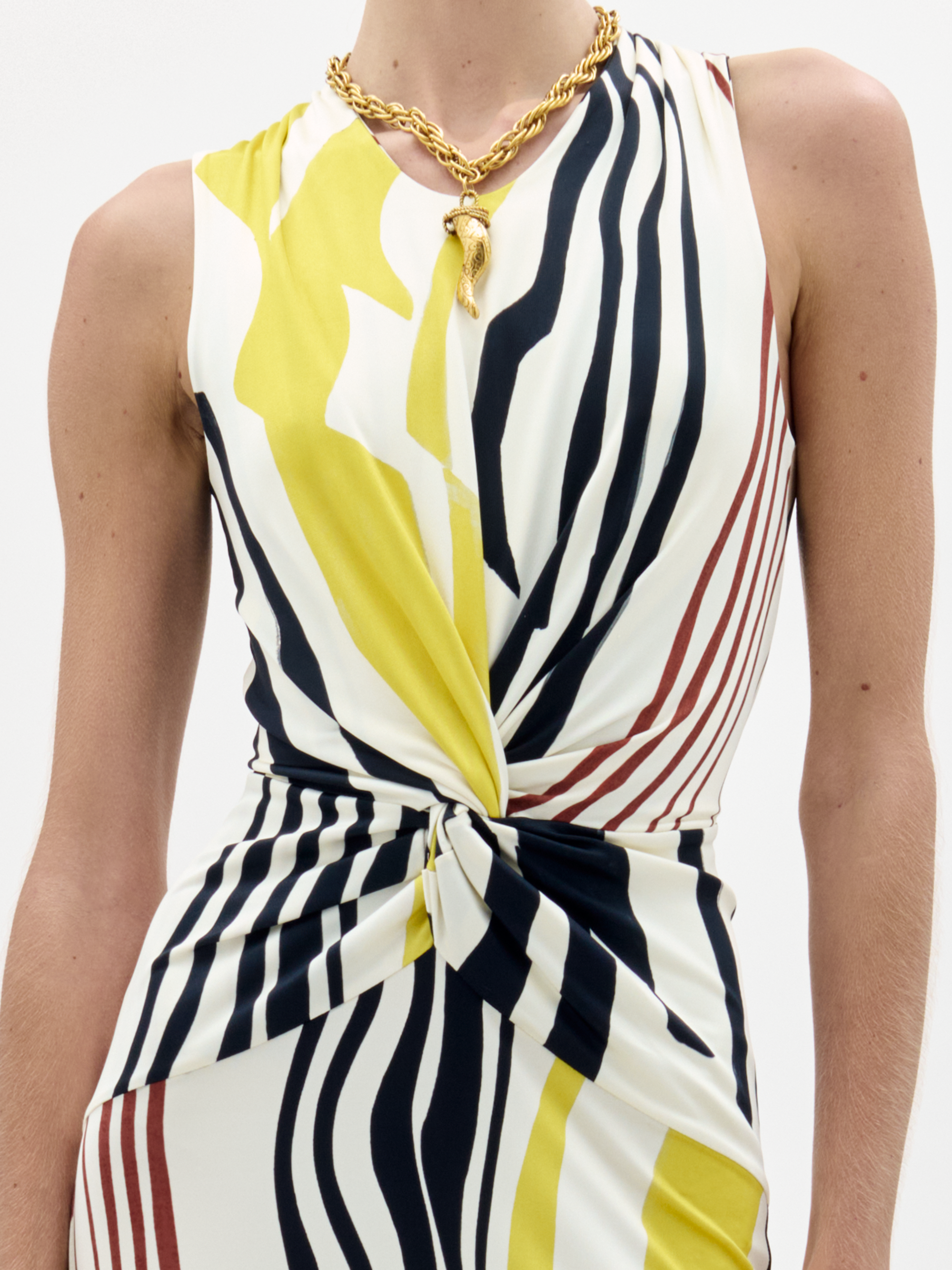 A woman stands against a white background wearing the Luna Dress—a sleeveless, ankle-length dress with yellow, black, and white vertical abstract stripes—and brown fringed shoes.